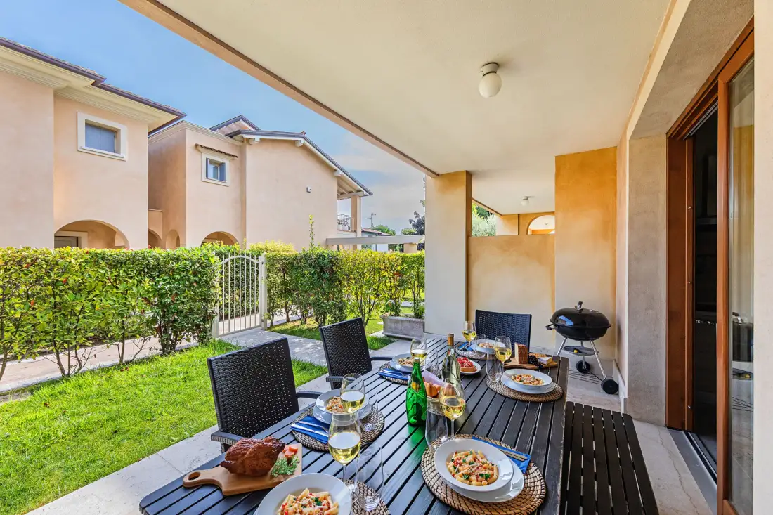 Covered patio with outdoor dining table and grill overlooking a hedge and lawn.