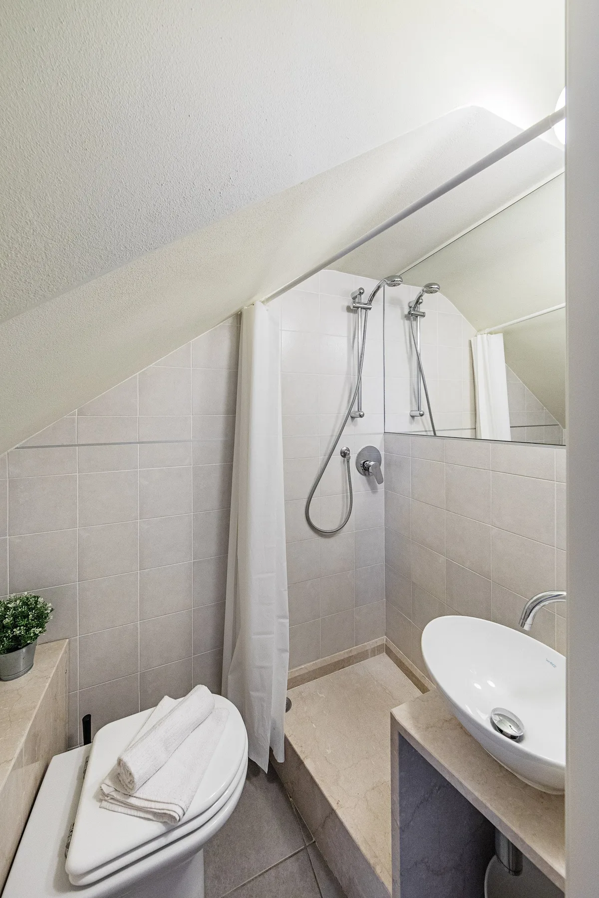 A compact bathroom with a shower, toilet, and sink beneath a sloped ceiling.