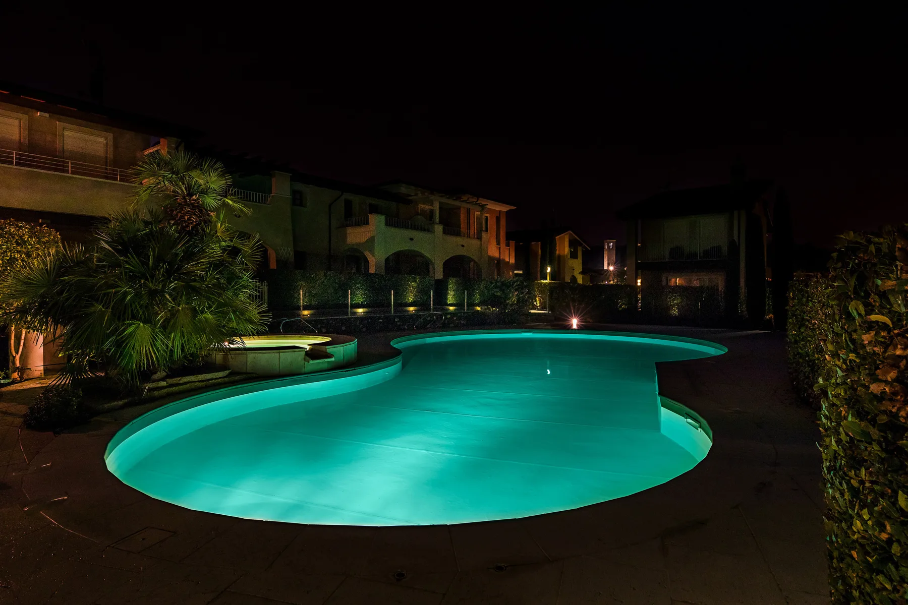 An illuminated swimming pool at night, surrounded by buildings and landscaping.