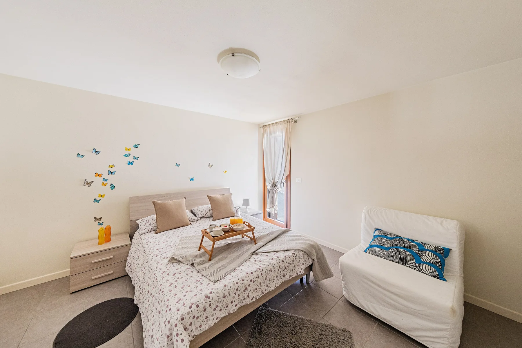 A bright bedroom with a double bed and a small armchair beside the window.