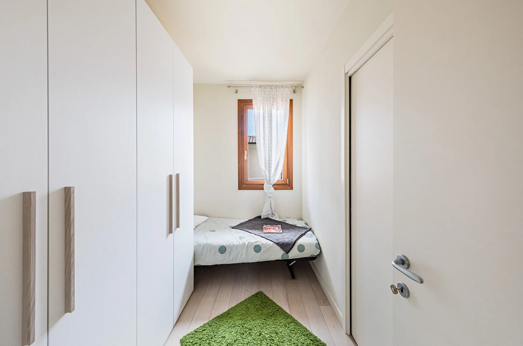 A narrow bedroom has a bed beneath a curtained window and a green rug.