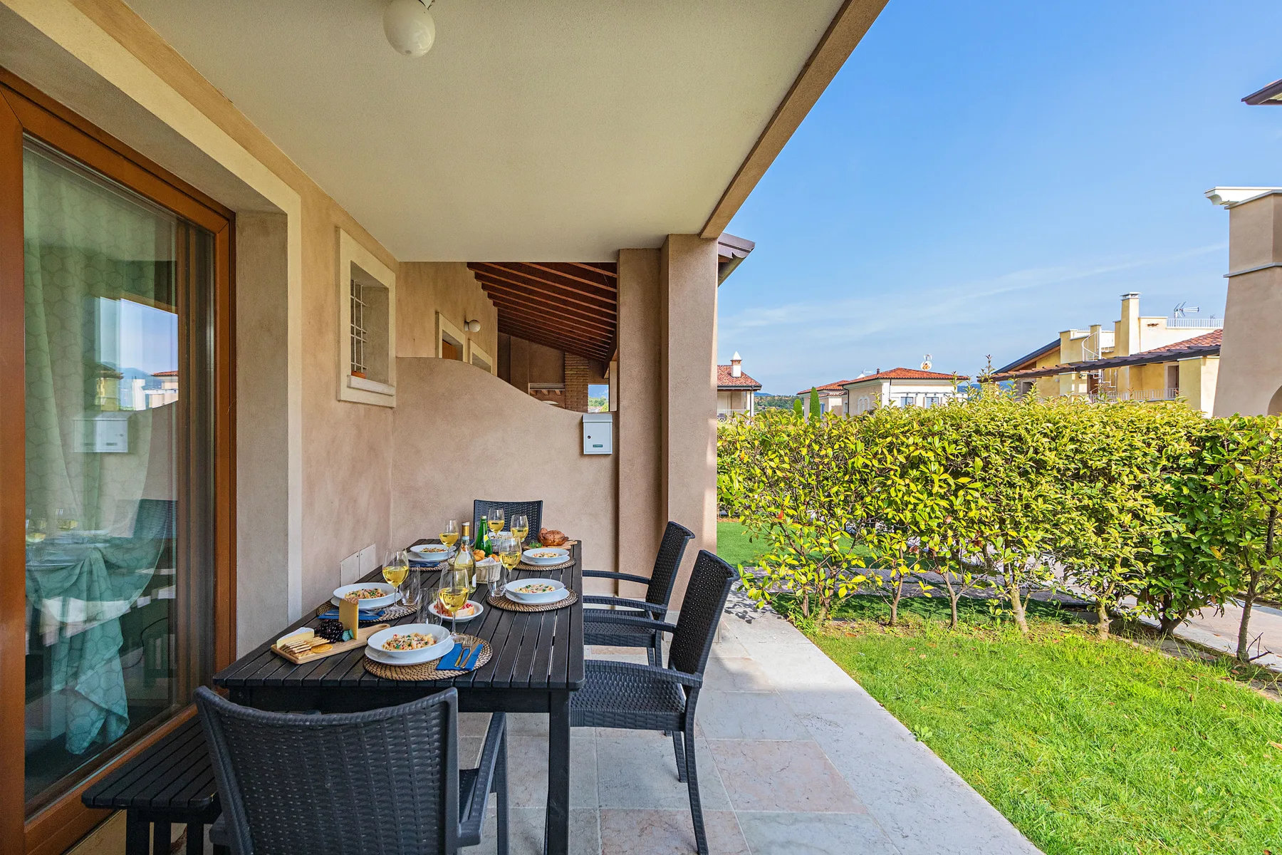 A covered outdoor dining area with a table beside a hedge and lawn.