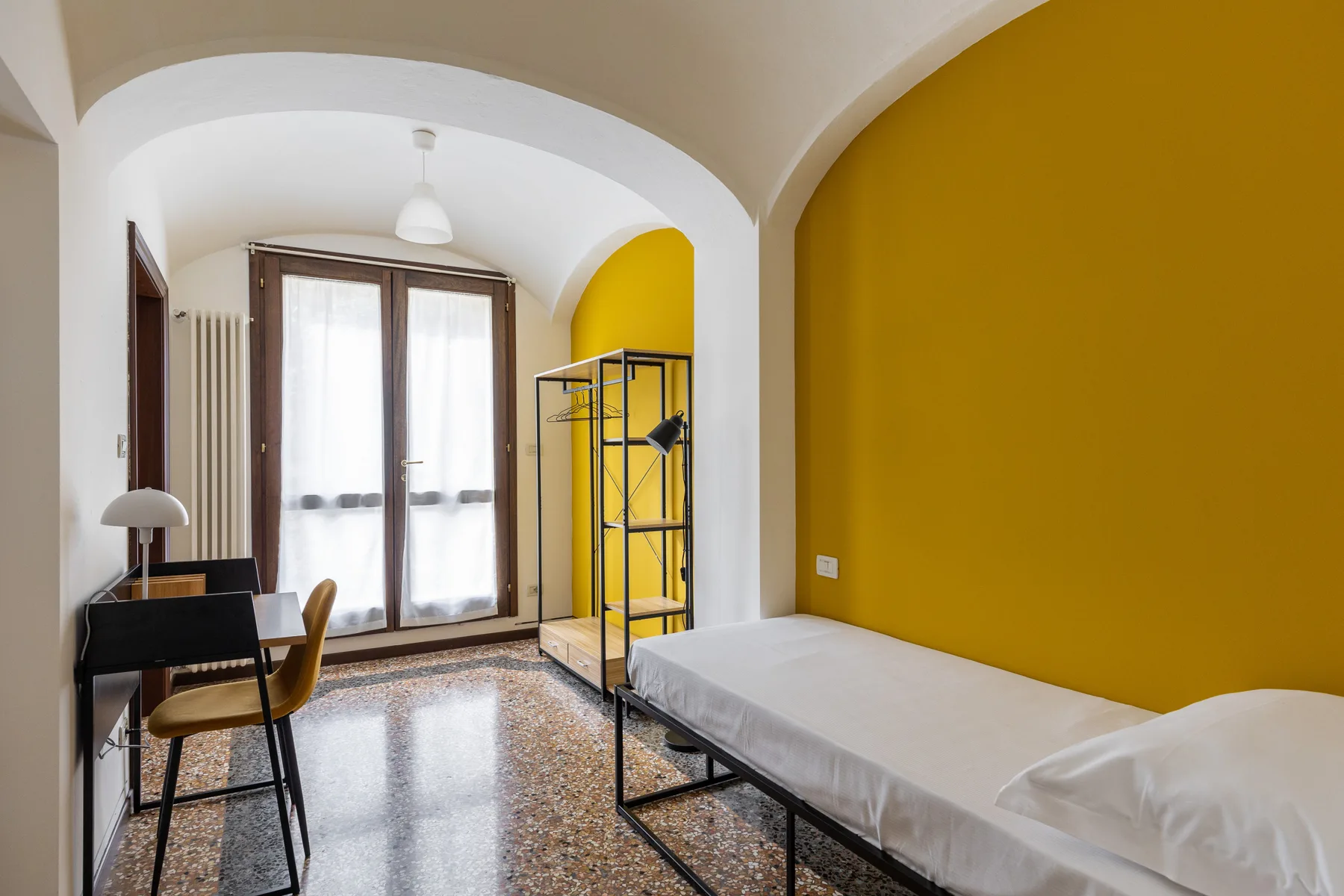 A bedroom with a bed, a desk, and tall windows.