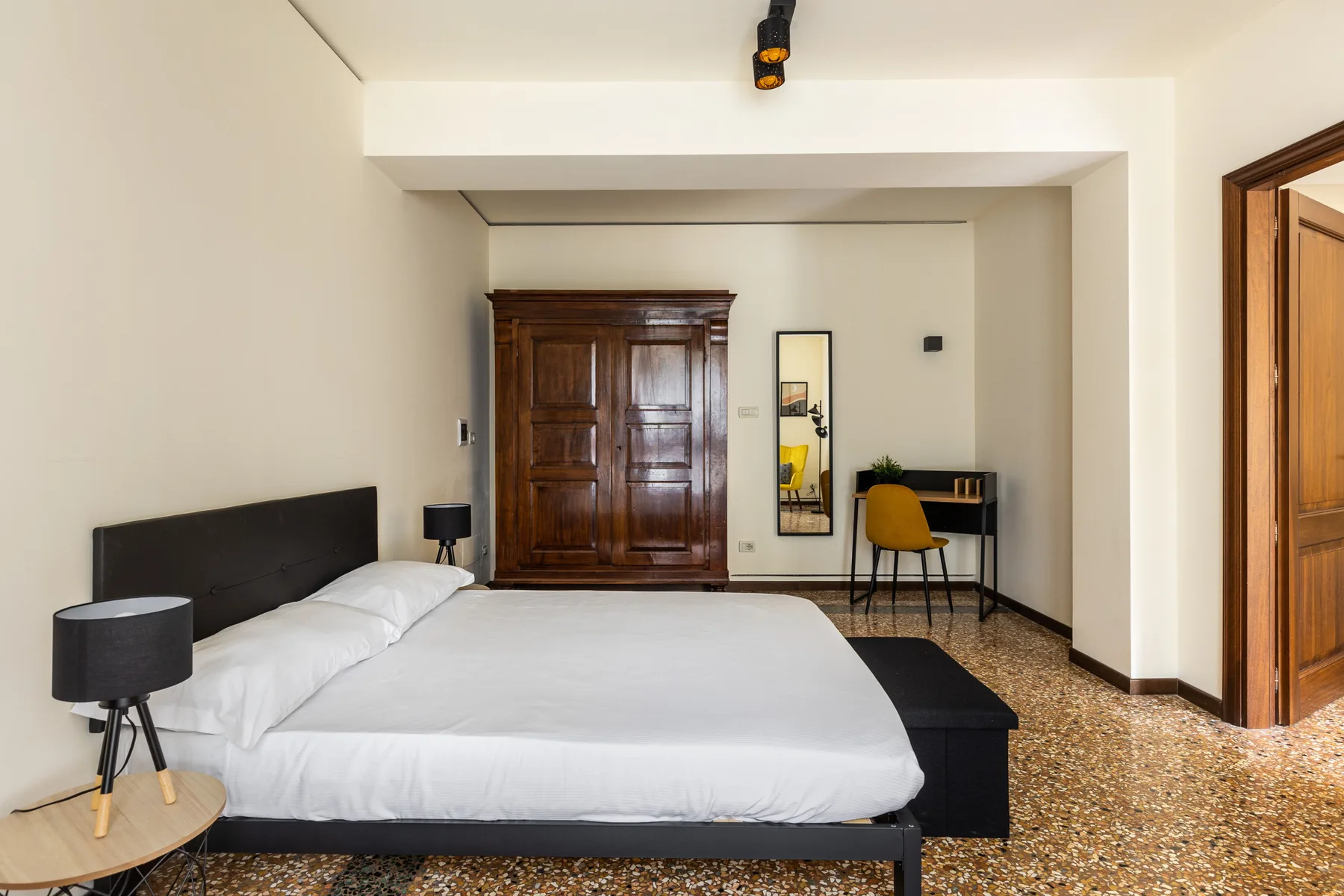 A bedroom with a large bed, a wooden wardrobe, and a small desk.