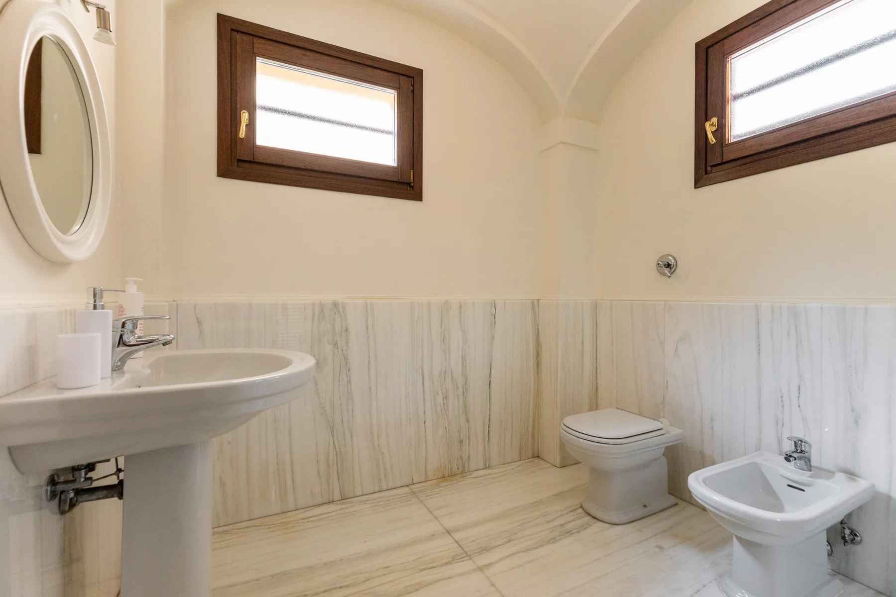 A bathroom with a sink, toilet, and bidet beneath high windows.