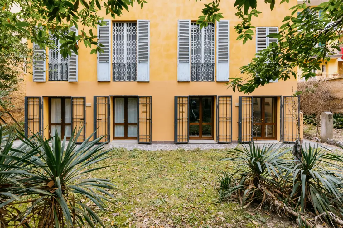 A yellow building faces a small garden with leafy plants and trees.