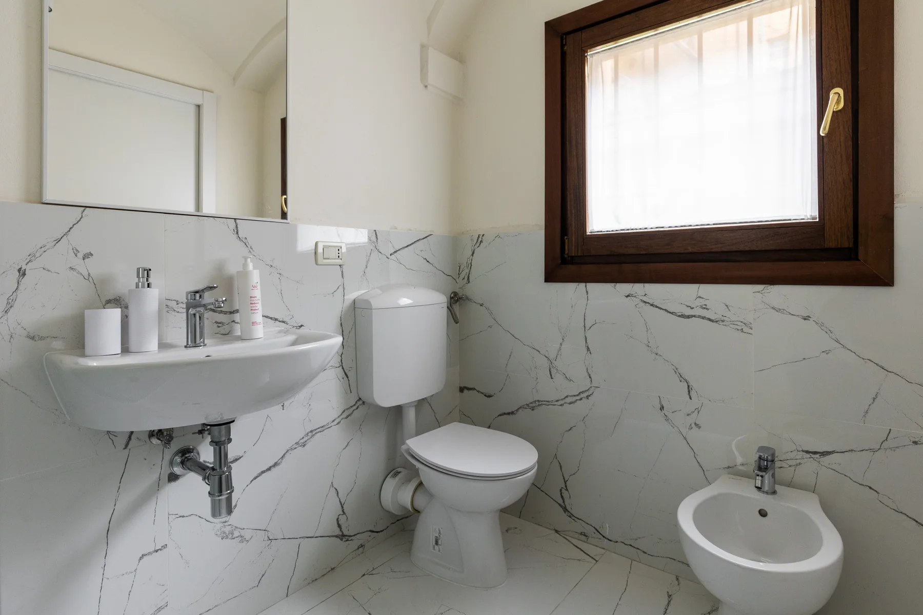 A bathroom with a wall-mounted sink, toilet, and bidet beneath a window.