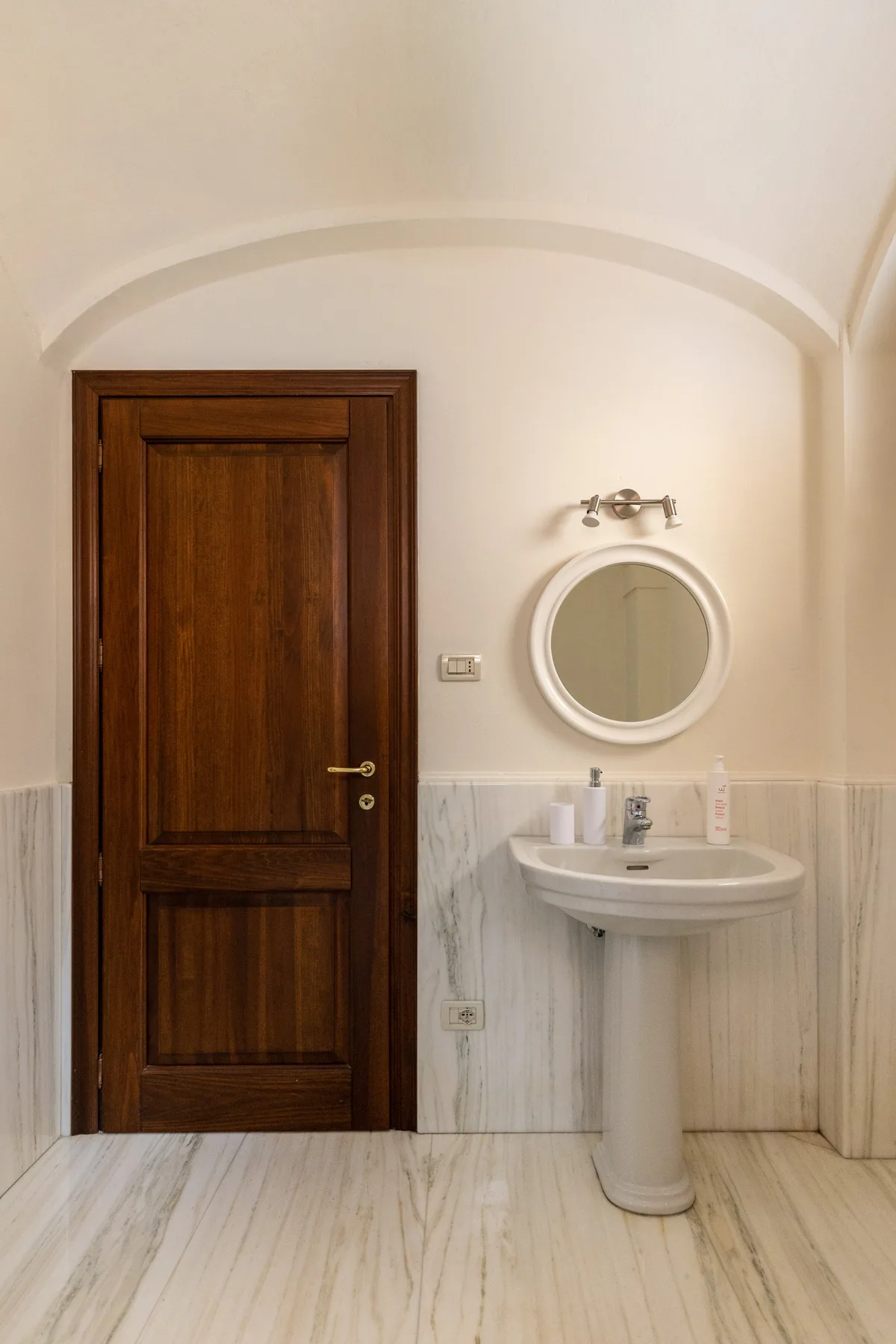 A bathroom with a pedestal sink, round mirror, and dark wooden door.