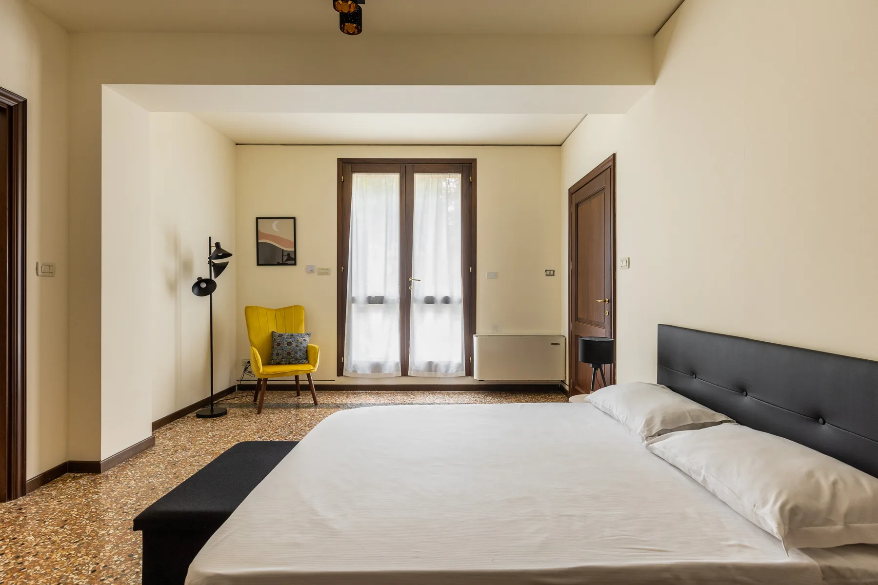 A bedroom with a large bed, yellow seating, and tall curtained windows.