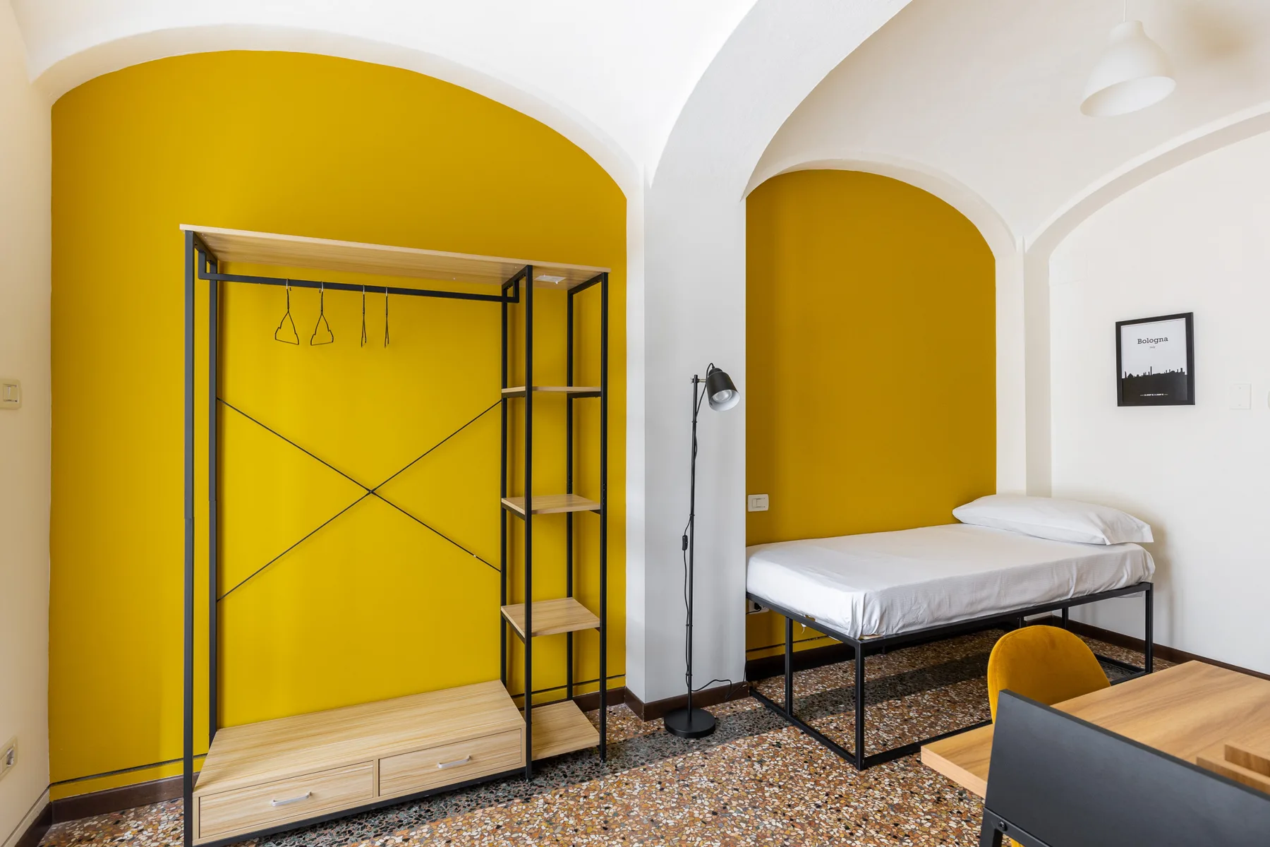 A bedroom with a bed beside an open clothes rack against a yellow wall.
