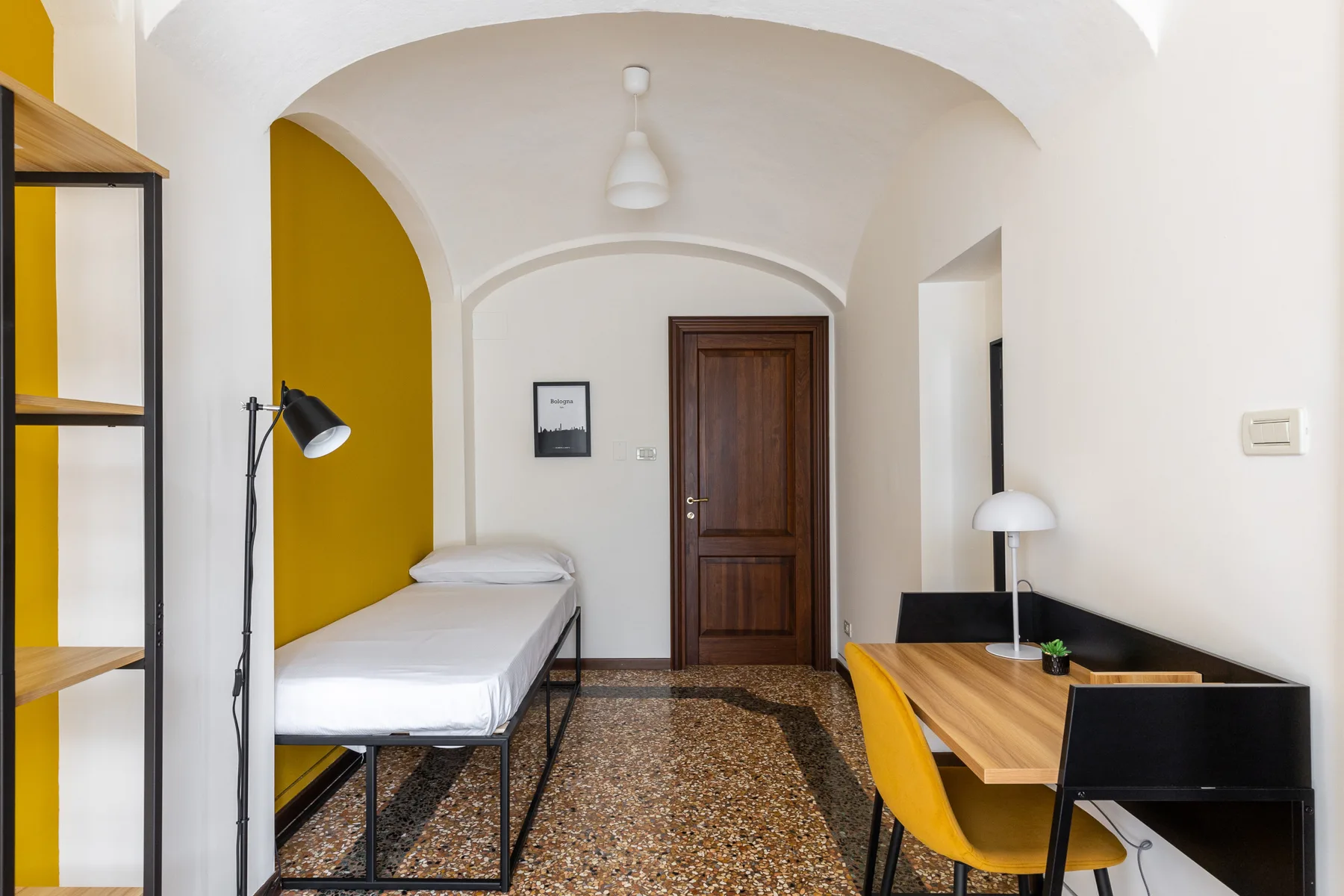 A compact bedroom with a single bed and a wooden desk beside a yellow wall.
