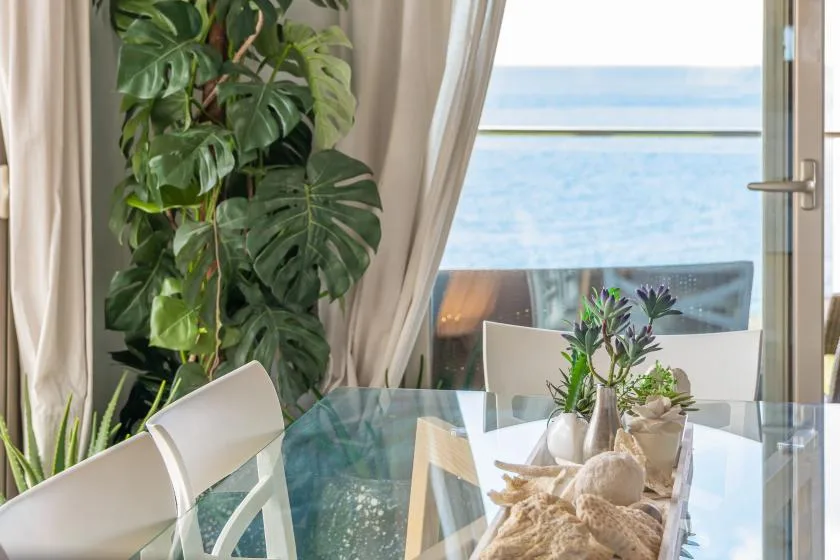 A glass dining table and chairs sit beside a bright window overlooking the water.