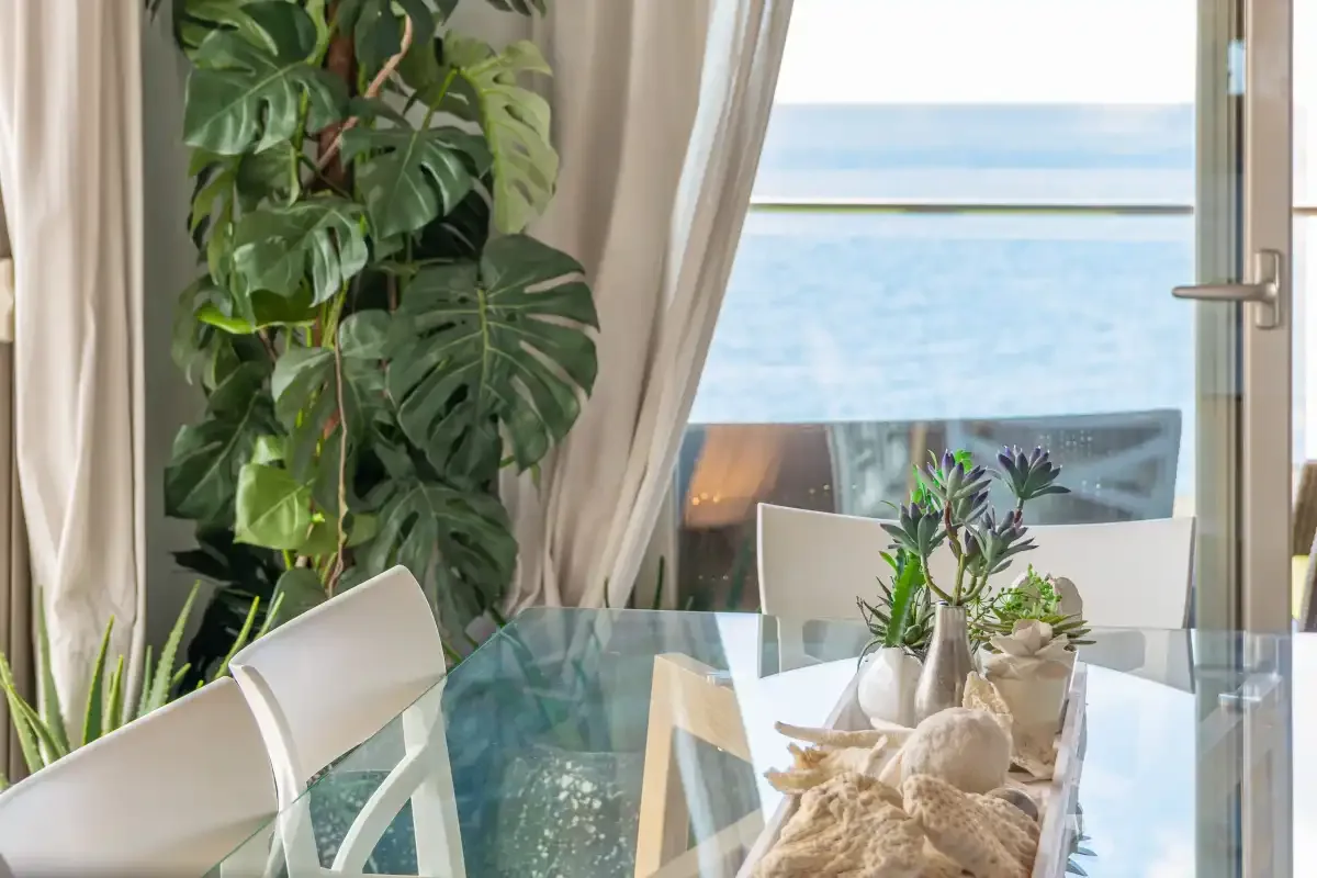 A dining area with a glass table and chairs faces the sea through open curtains.