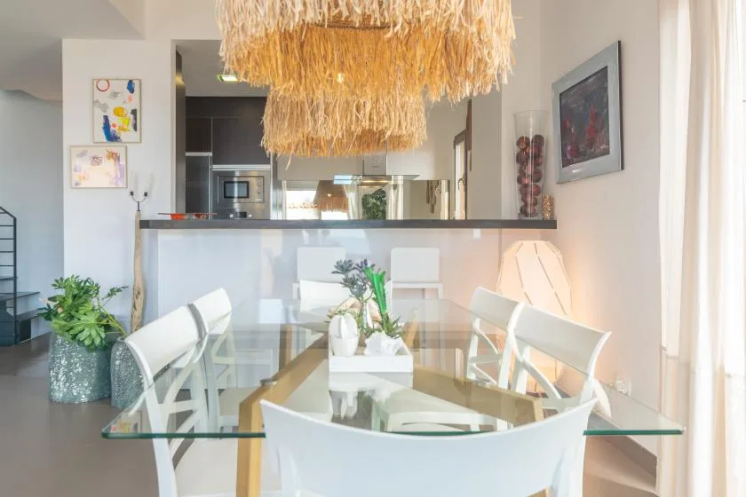 A dining area with a glass table, white seating, and a large hanging light.