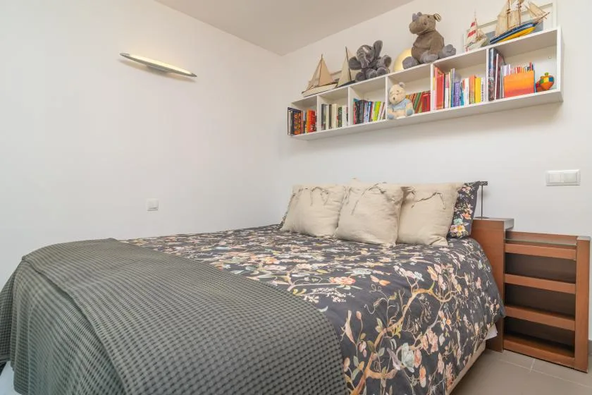 A bedroom with a patterned bedspread and a wall shelf of books and toys.