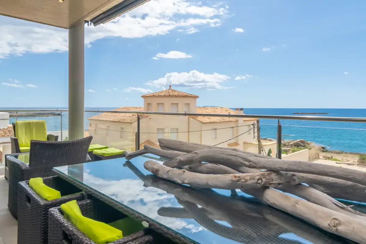 A covered terrace has dark seating and a glass table overlooking the sea.