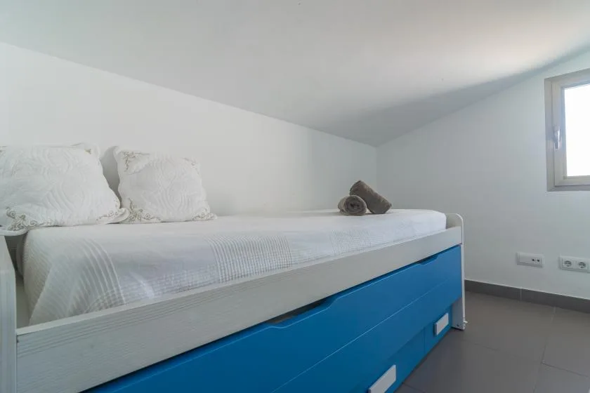 A low-ceiling bedroom with a white bed above blue storage drawers.