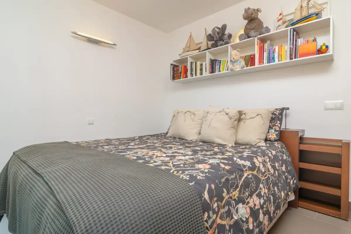 A bedroom with a bed and a wall shelf holding books and toys.