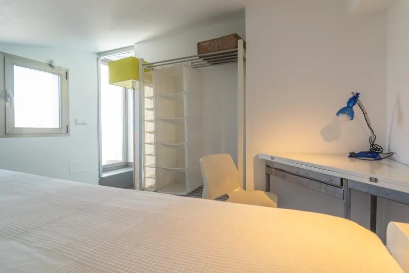 A bright bedroom with a bed, shelving, and a desk with a blue lamp.