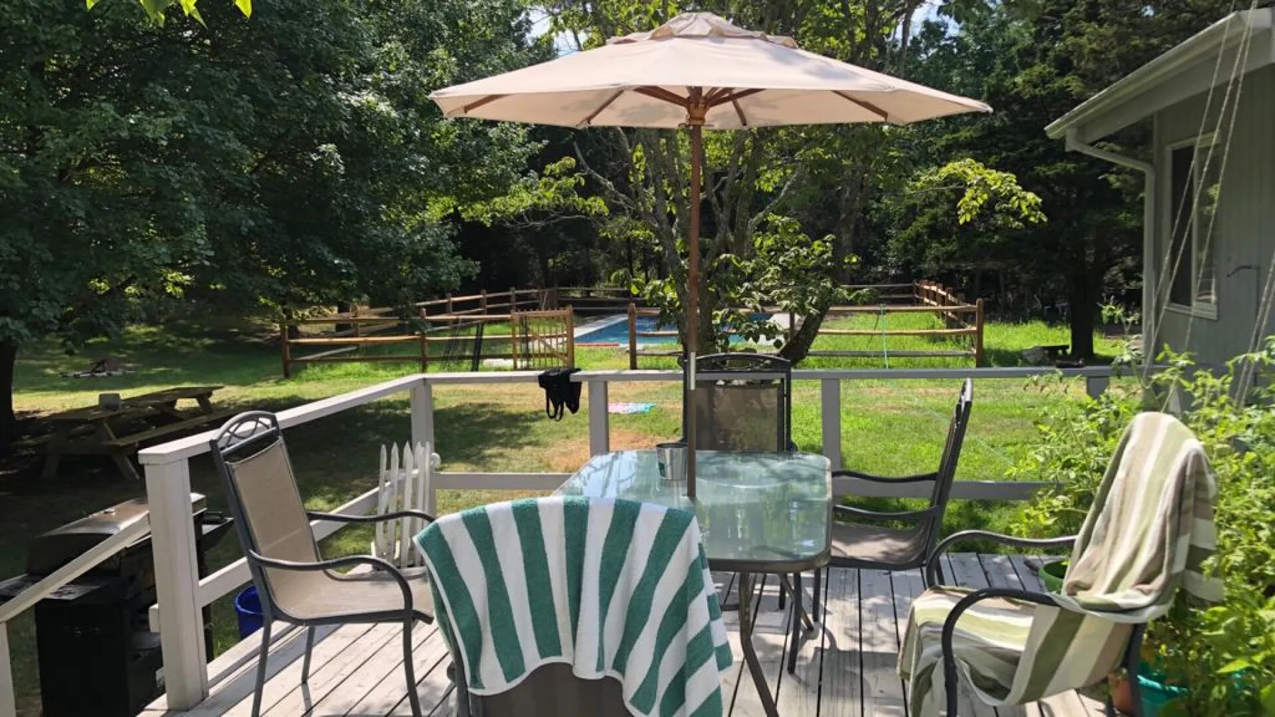 A patio seating area with a striped towel and umbrella overlooks a fenced yard.