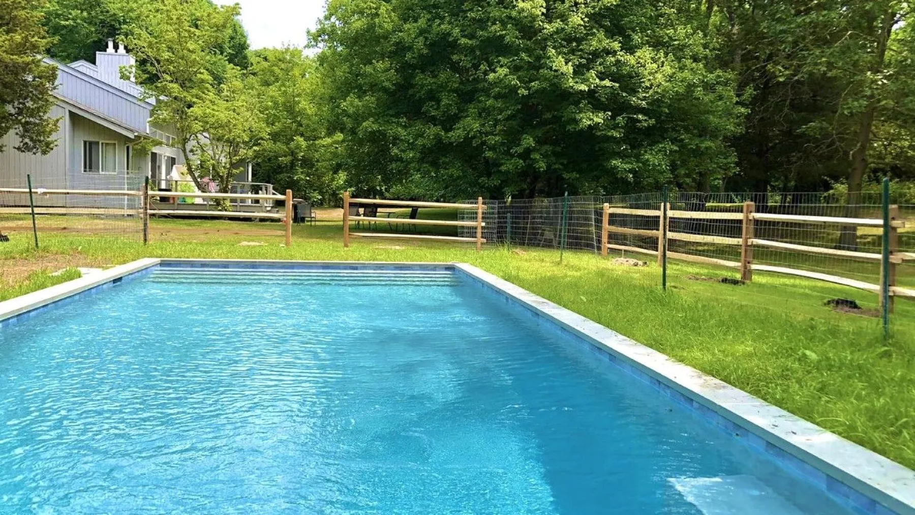 An outdoor swimming pool sits beside a grassy yard and leafy trees.