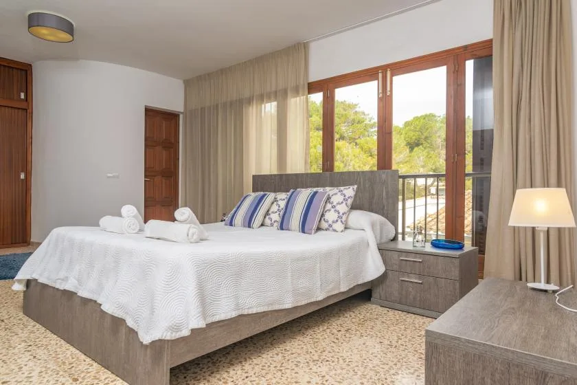 A bedroom with a neatly made bed and large windows overlooking green trees.