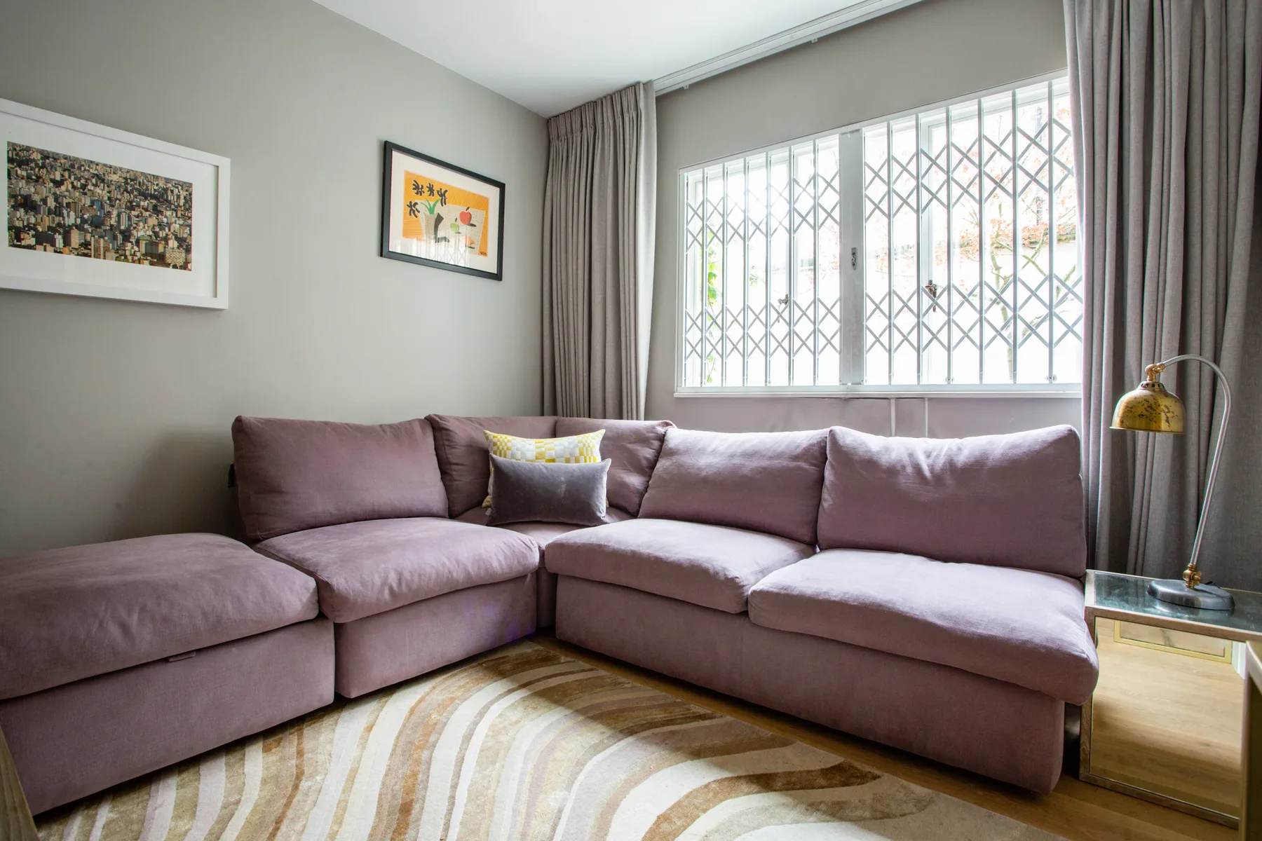 A living room with a mauve sectional and a large curtained window.