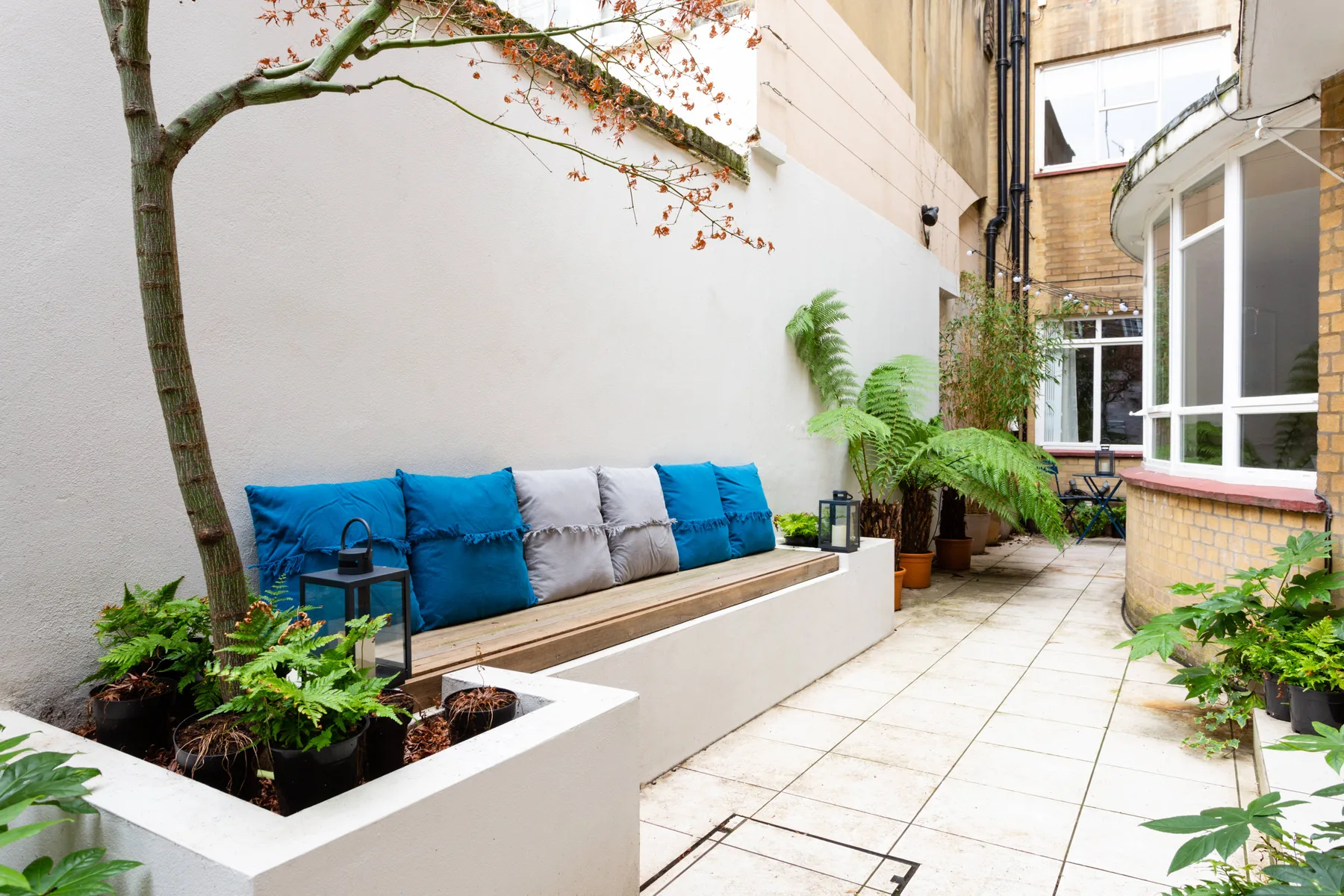 A tiled courtyard patio has a built-in bench with colorful cushions and surrounding plants.