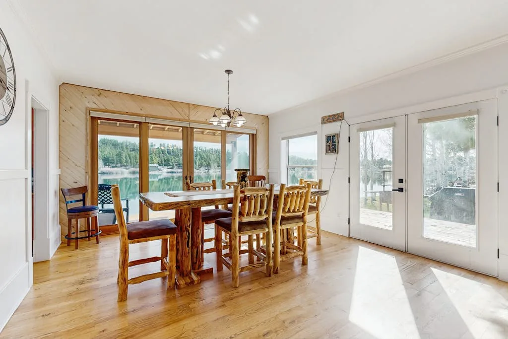 Un comedor luminoso tiene una mesa larga de madera junto a grandes ventanas y puertas de vidrio.