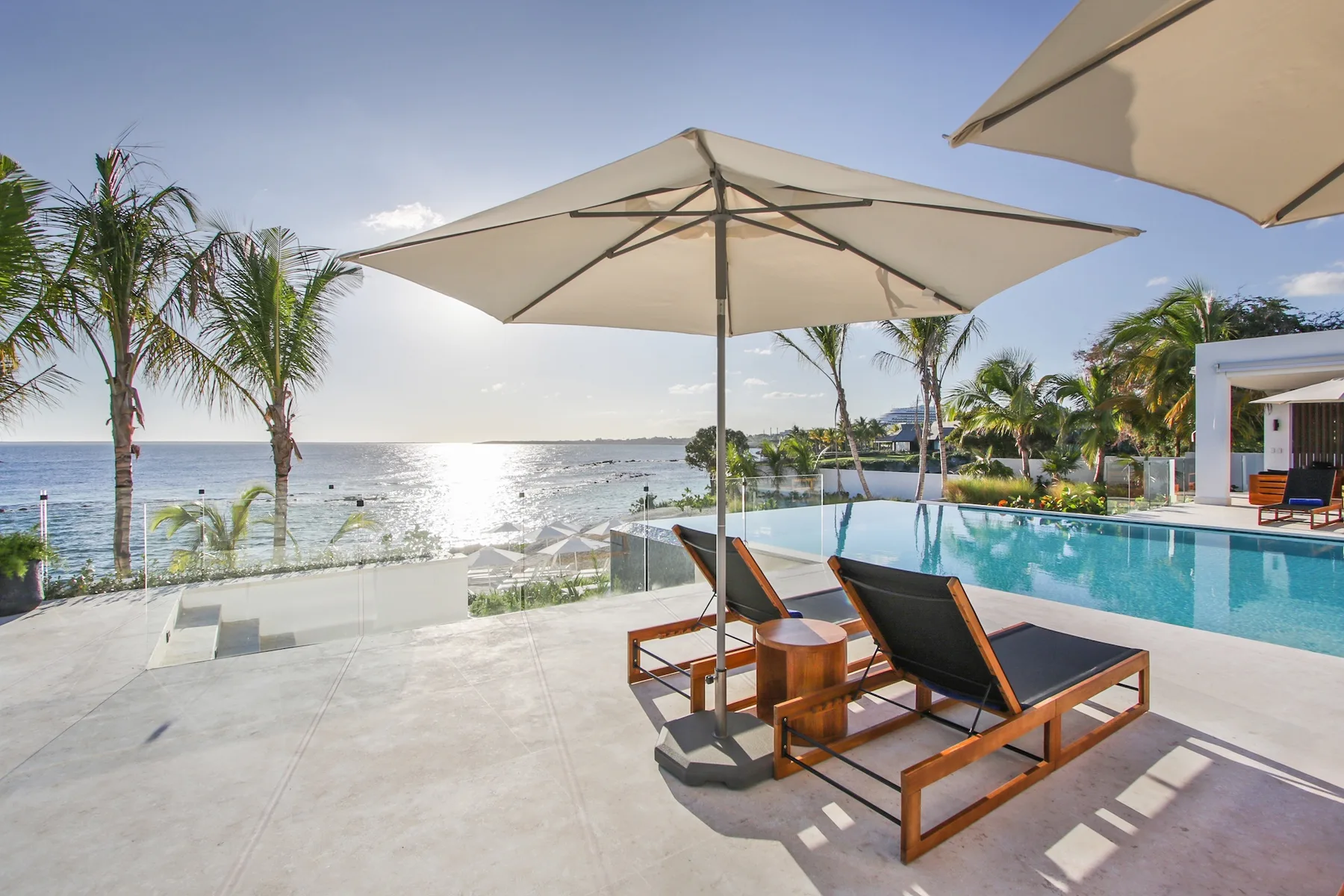 A poolside terrace overlooks the ocean, with lounge seating beneath a large umbrella.