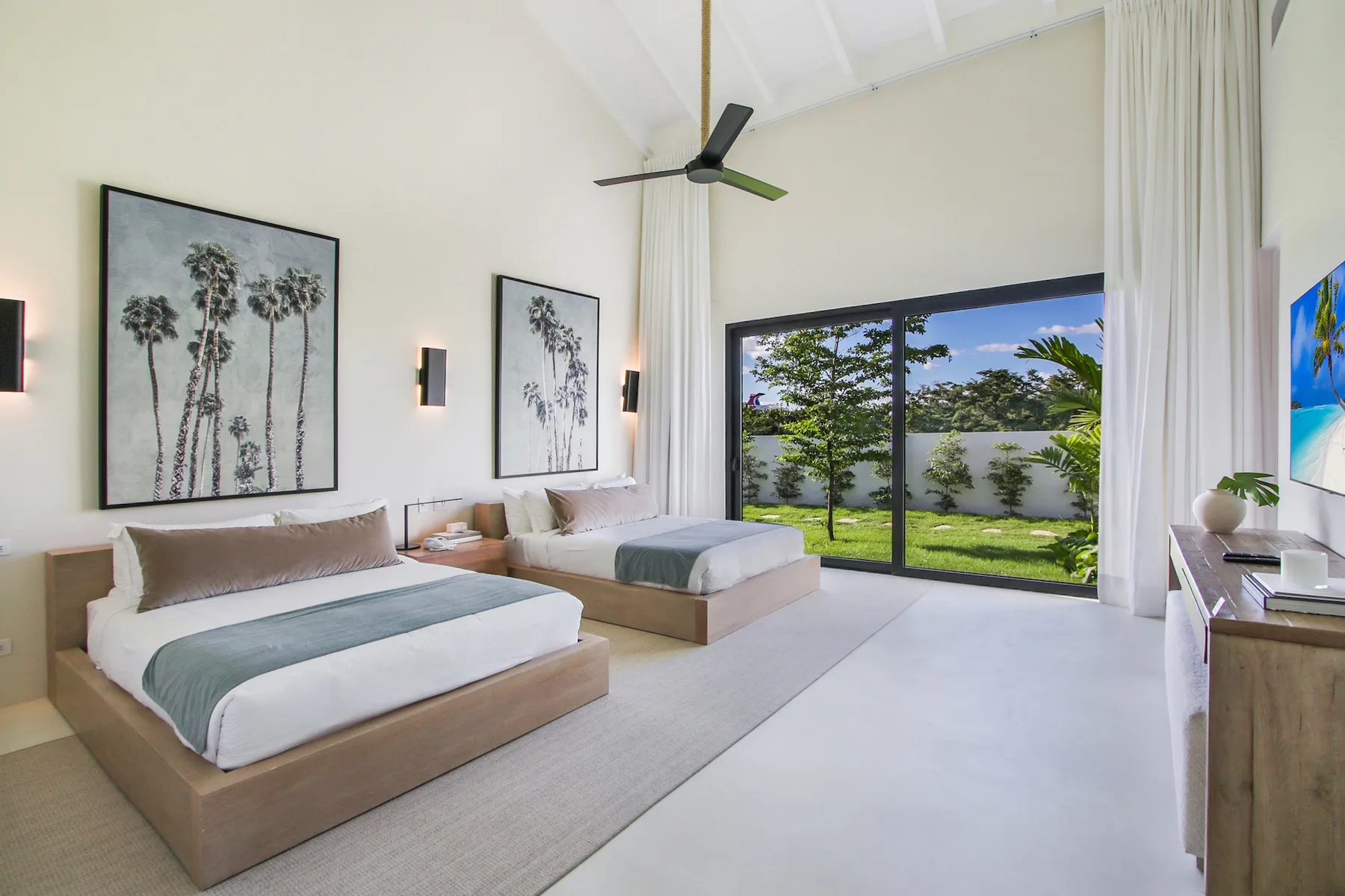 A bright bedroom with two beds beside a large window overlooking greenery.