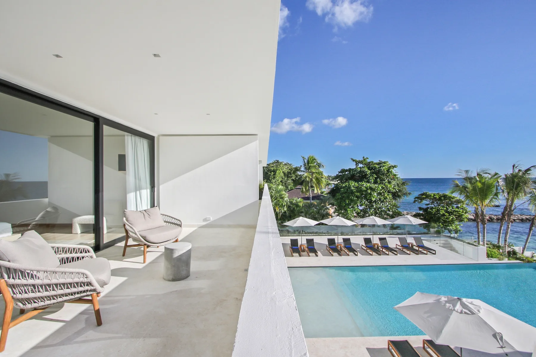 A modern balcony overlooks a swimming pool, palm trees, and the ocean.