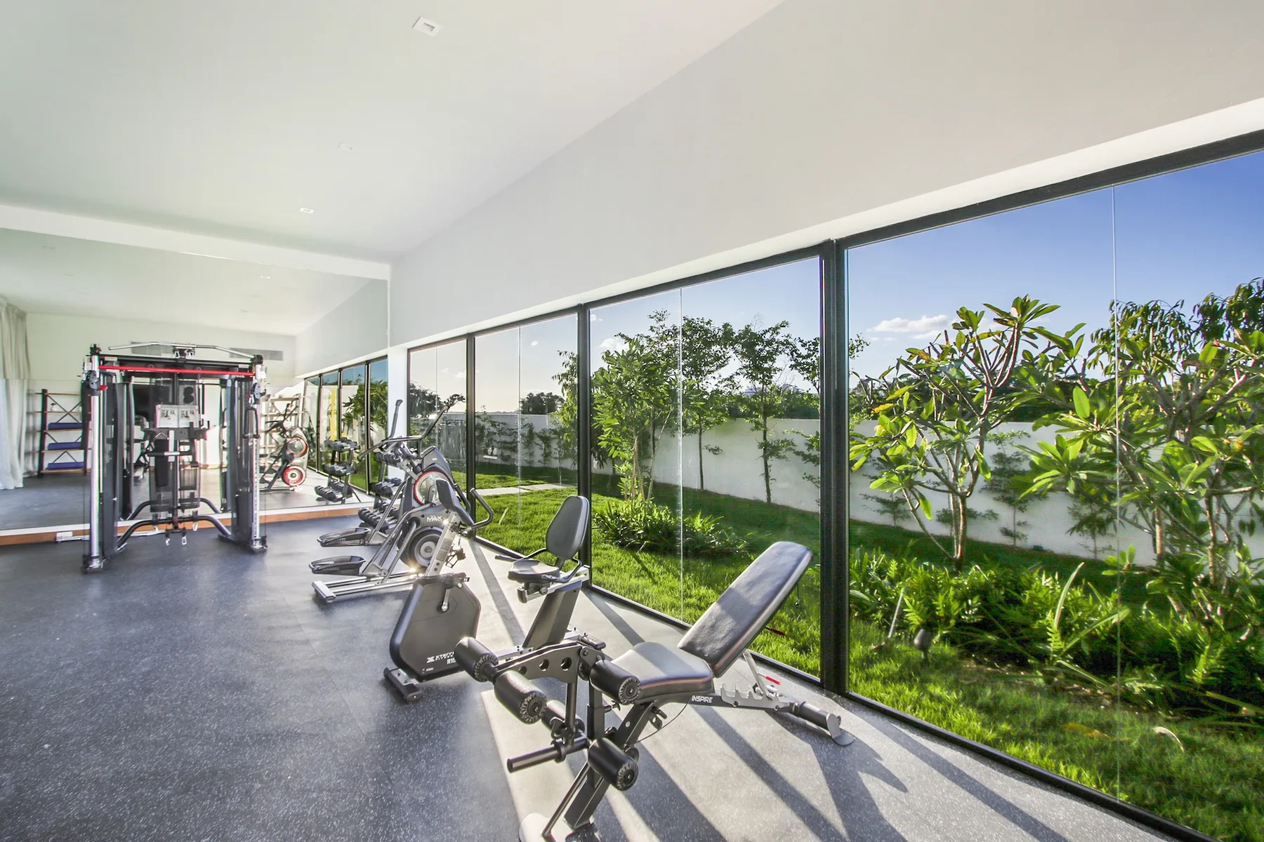 A bright indoor gym with exercise machines beside large windows overlooking greenery.