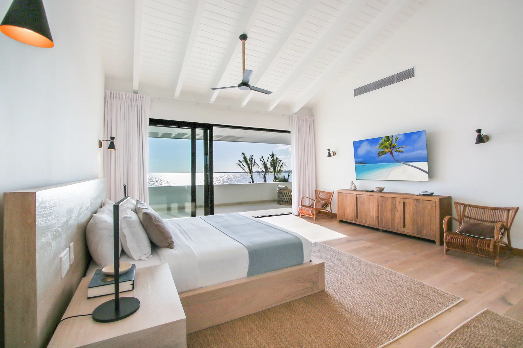 A bright bedroom with a bed, ceiling fan, and sliding glass doors overlooking the sea.