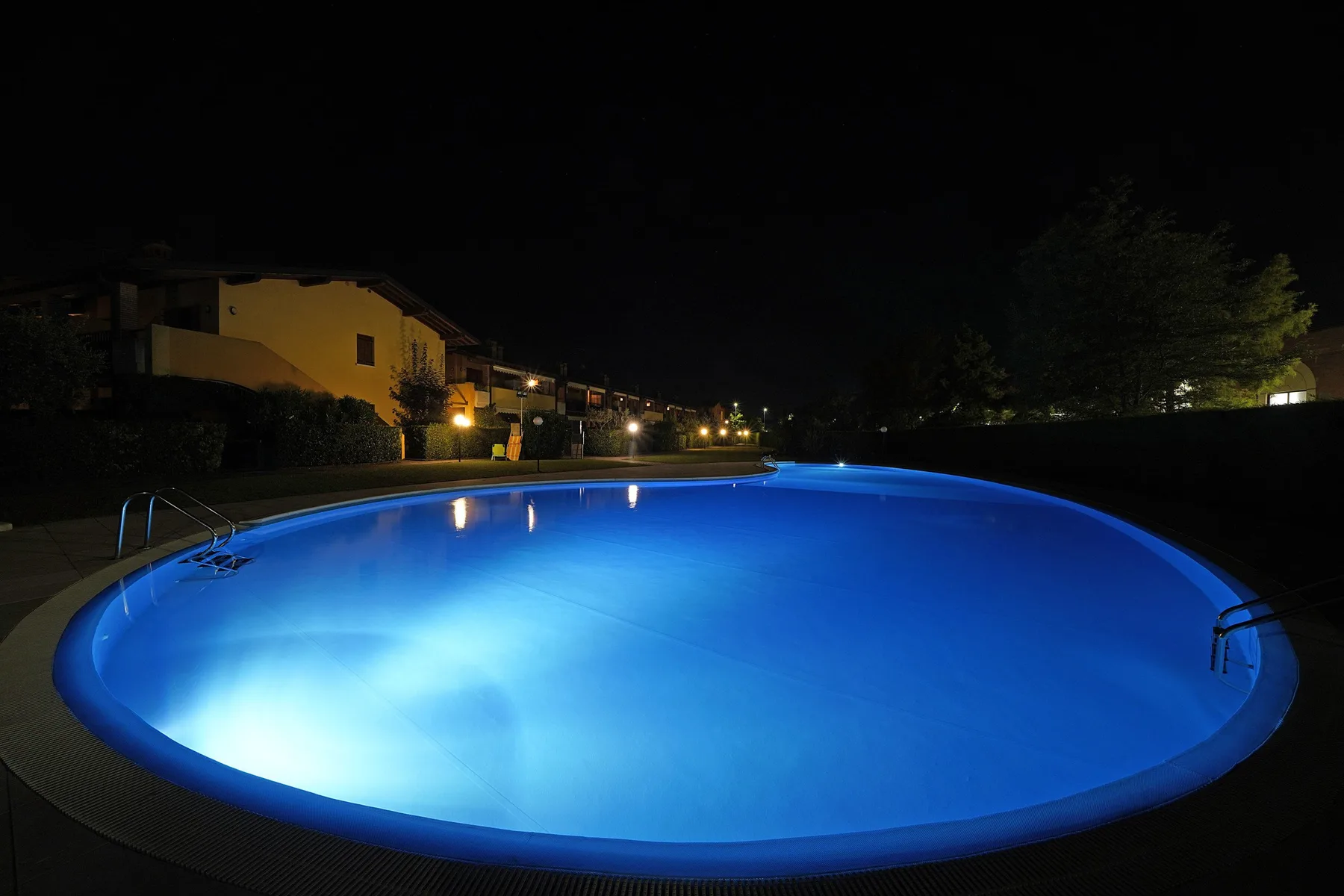 A swimming pool glows blue at night beside illuminated buildings.