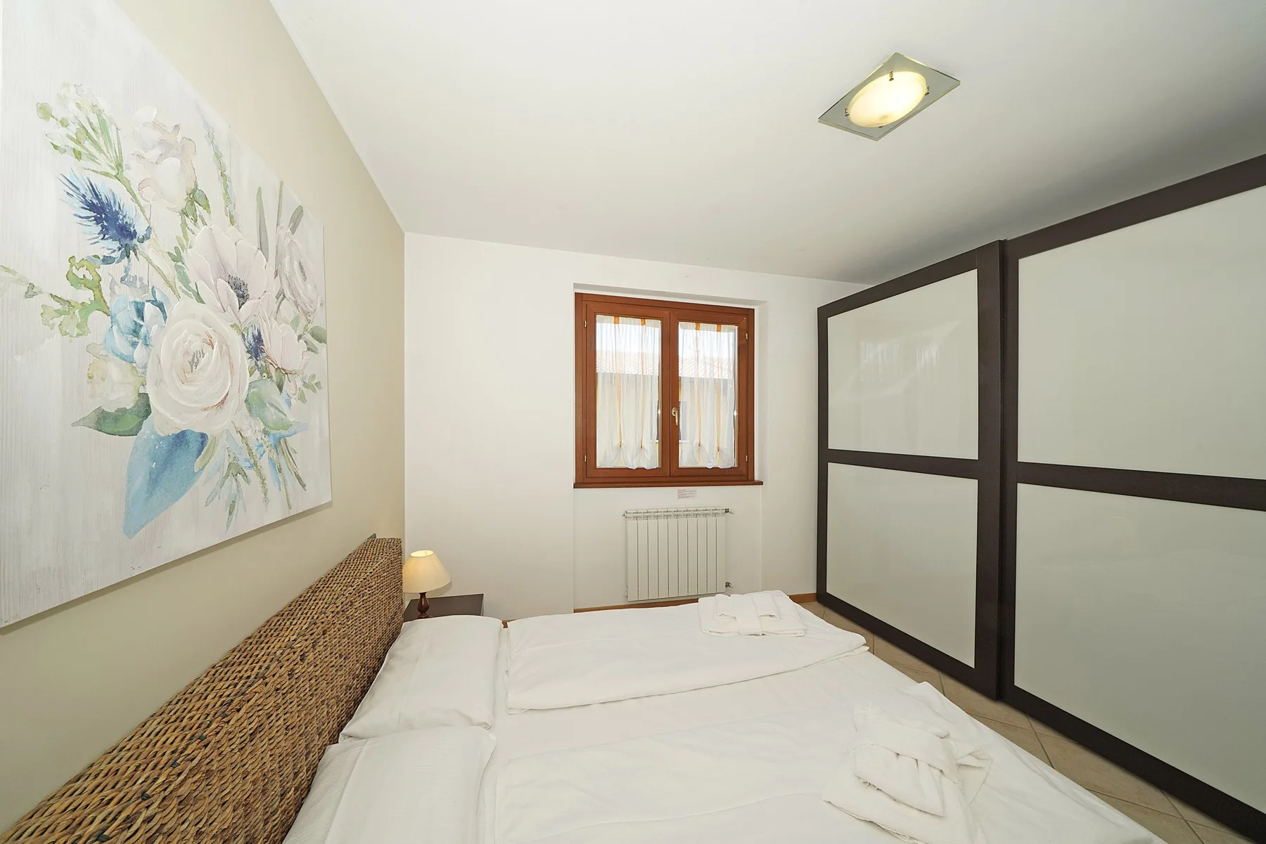 A bedroom with a white bed, floral wall art, and a window.