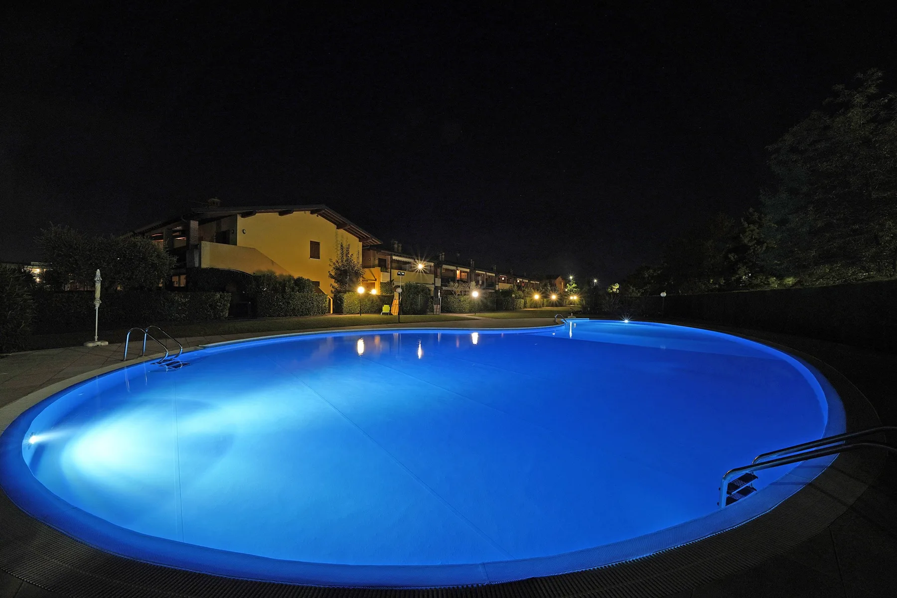 A brightly illuminated swimming pool at night, with buildings and trees in the background.