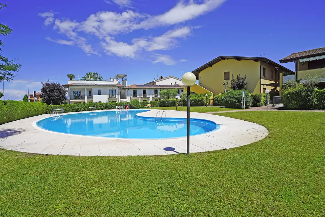An outdoor swimming pool sits beside a grassy lawn and low buildings.