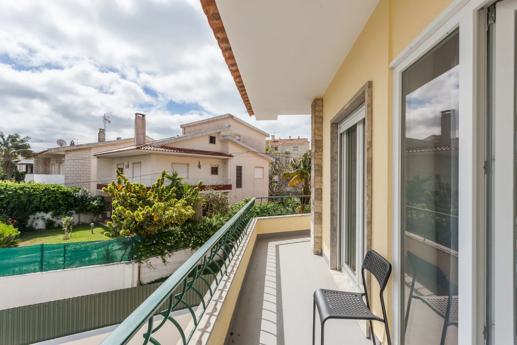 Un balcón con asientos metálicos da a un jardín y casas cercanas.