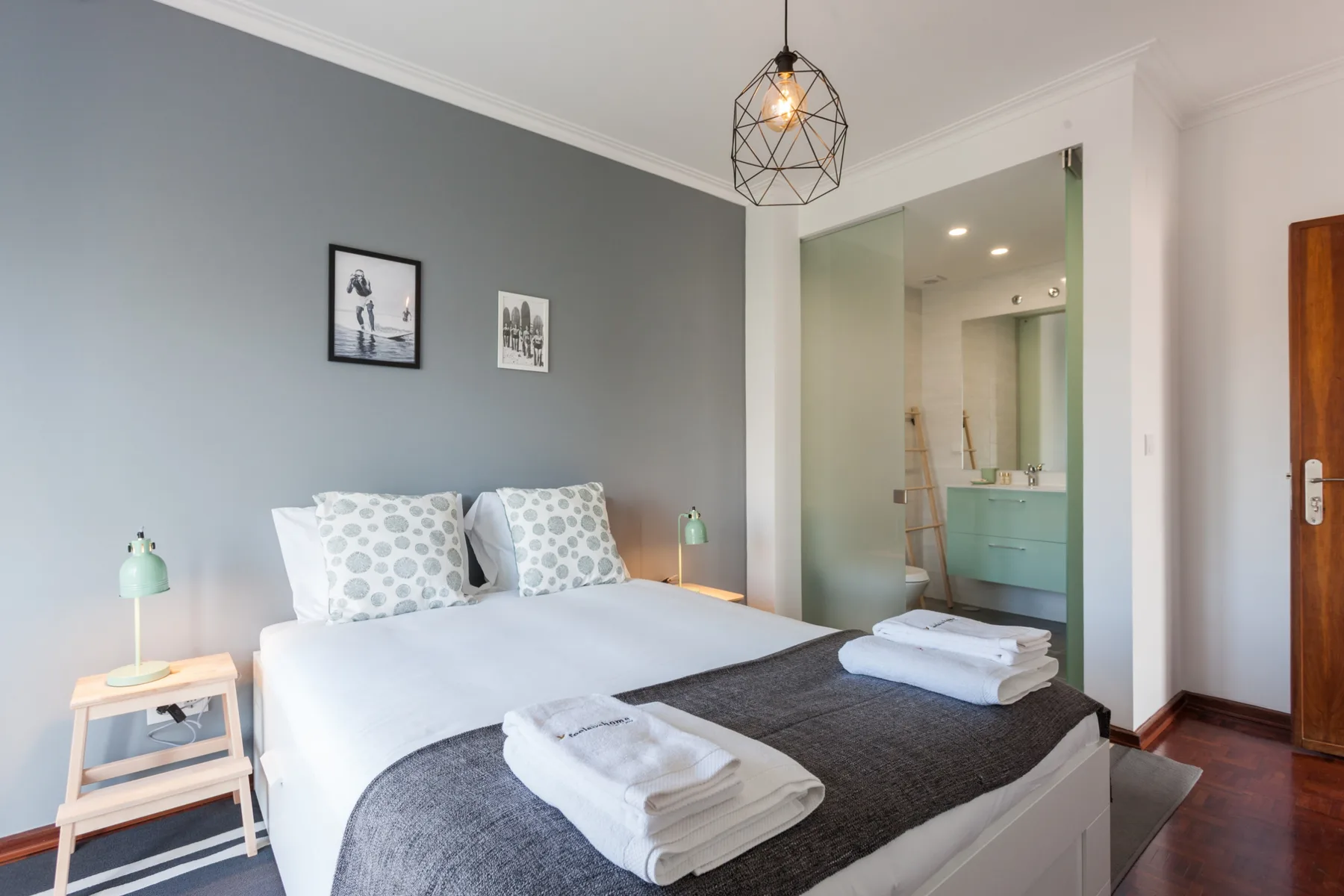 Un dormitorio con una cama vestida con ropa de cama blanca y gris, junto a una zona de baño abierta.