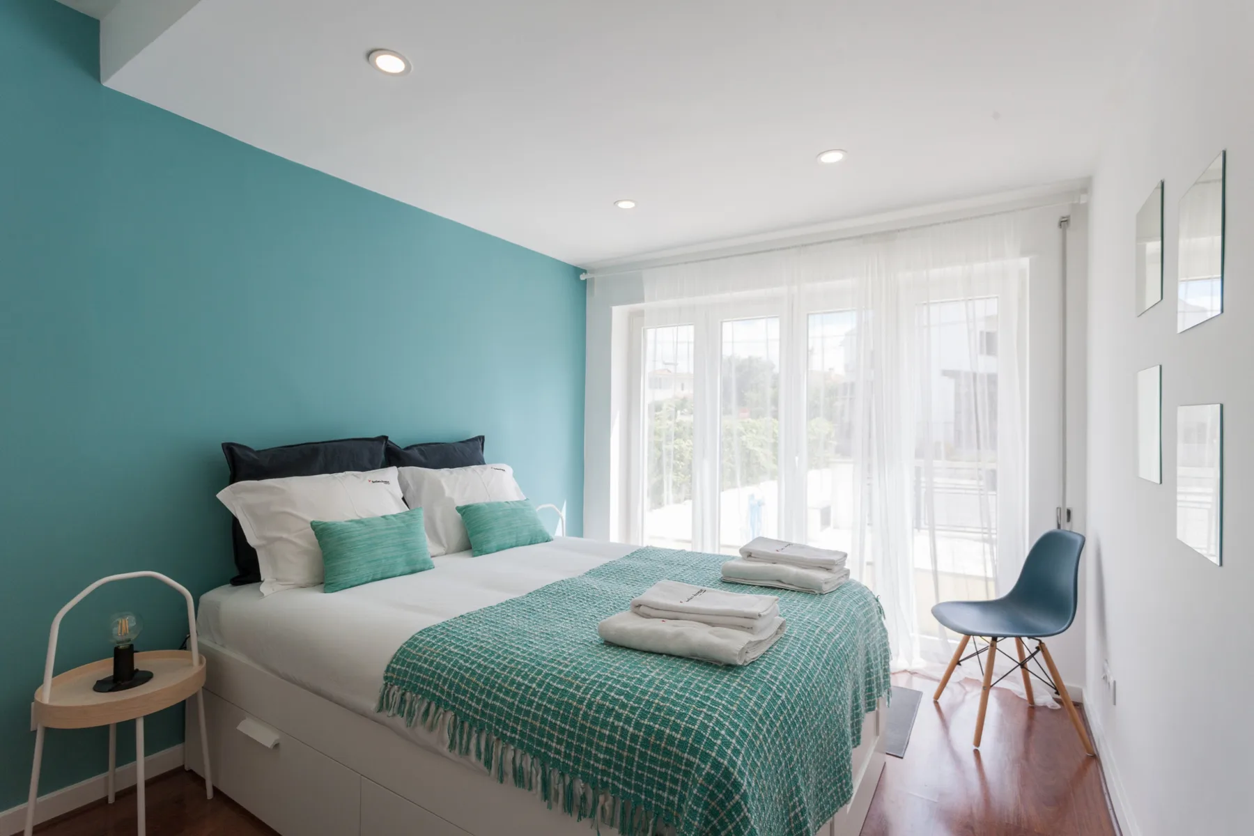 Un dormitorio con una cama vestida de blanco y turquesa junto a una ventana luminosa.
