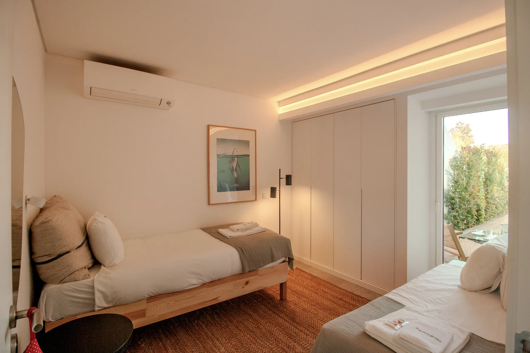 A bright bedroom with two beds and a glass door opening onto a balcony.