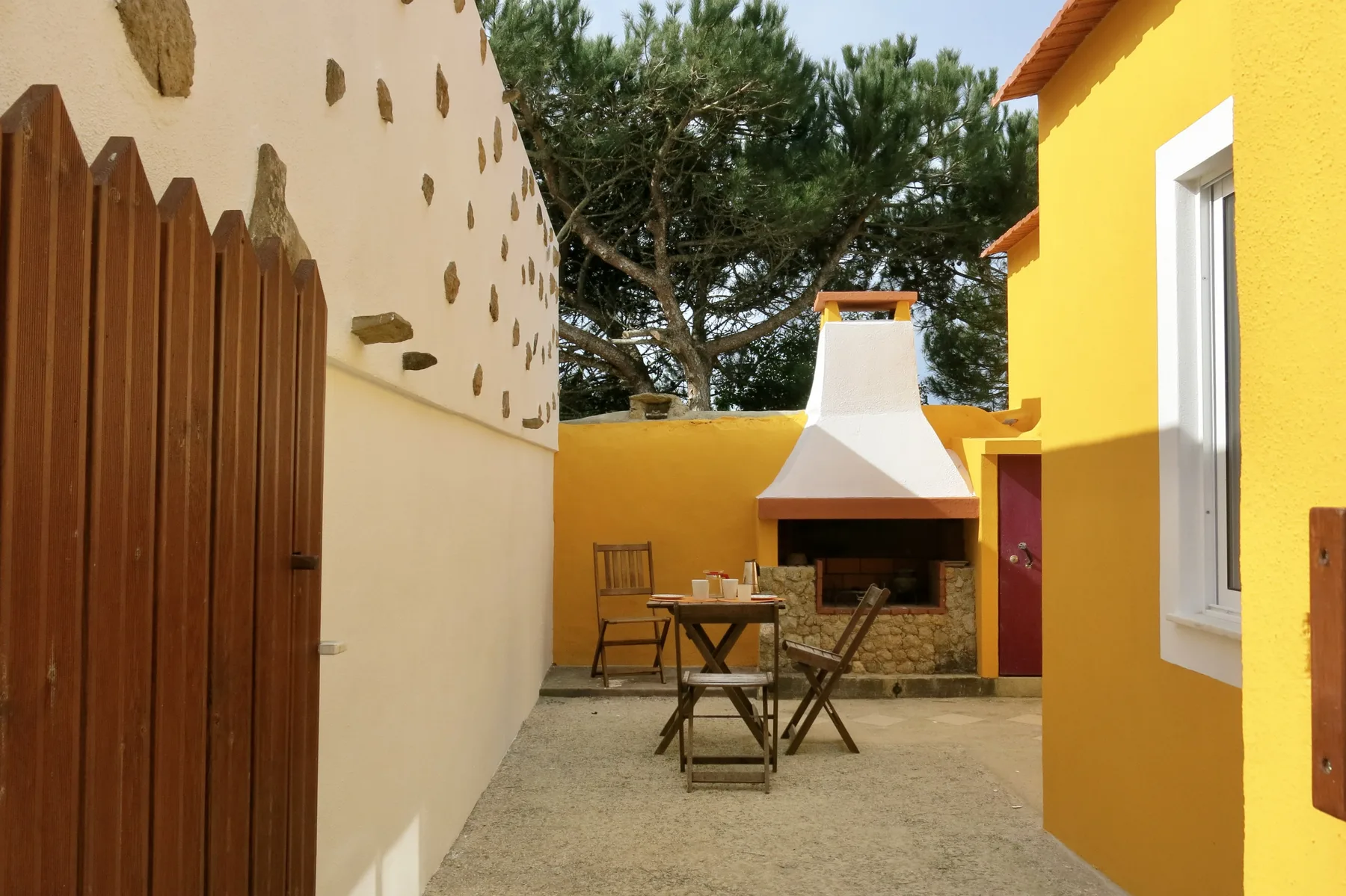 A sunny courtyard with a wooden dining table beside an outdoor fireplace.
