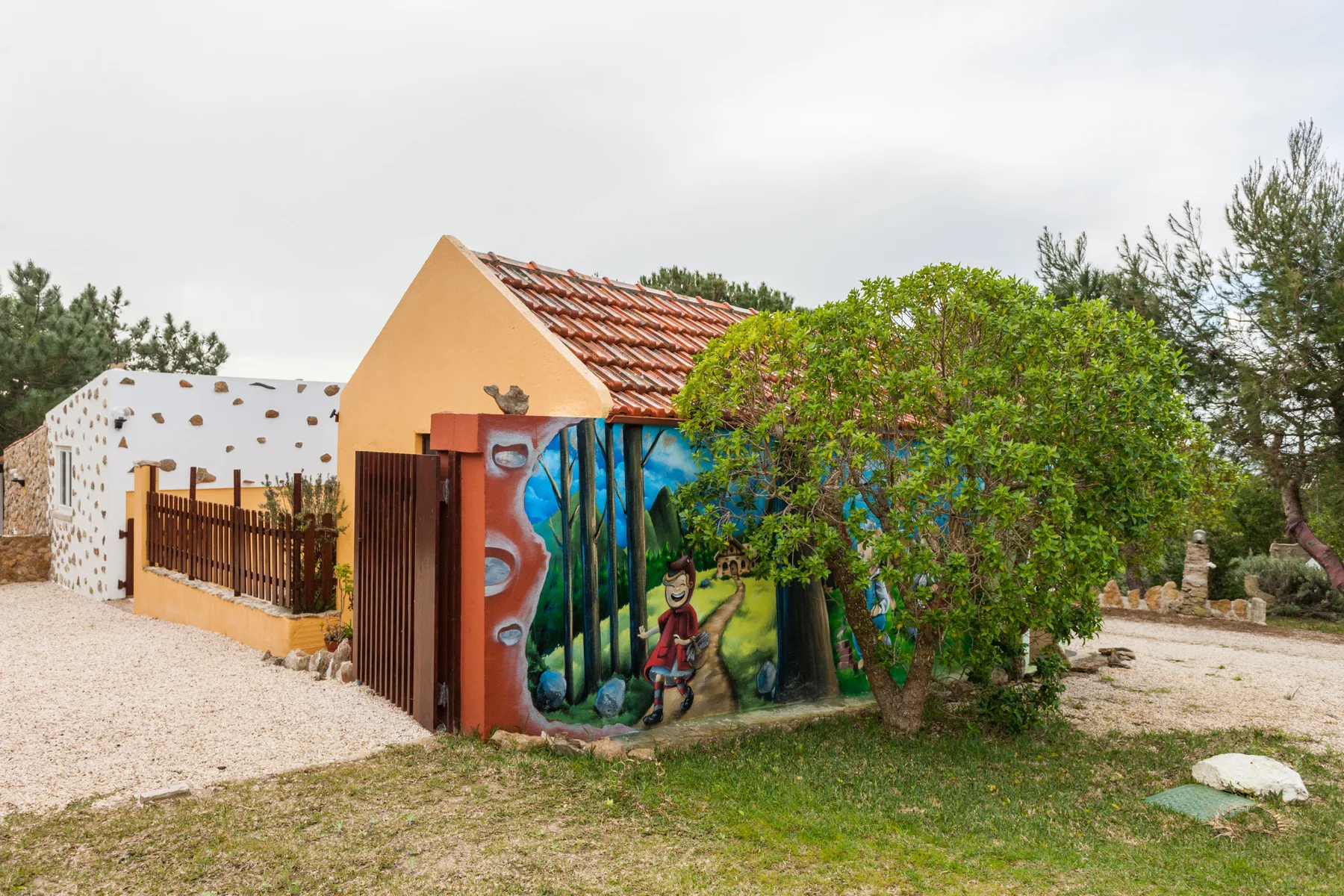A low house with a colorful mural stands beside a gravel yard and leafy trees.