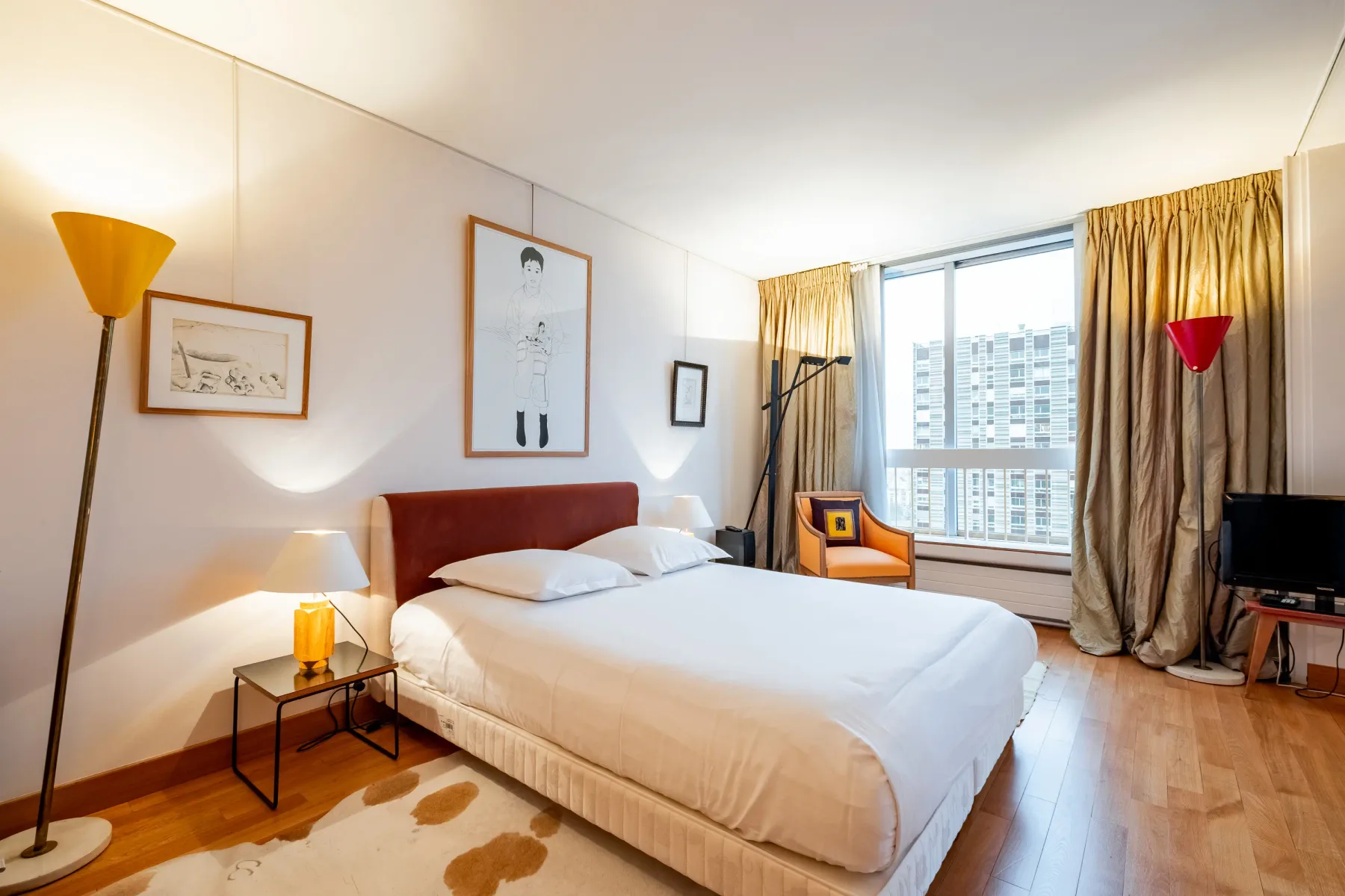 A bedroom with a neatly made bed, artwork, and a large curtained window.