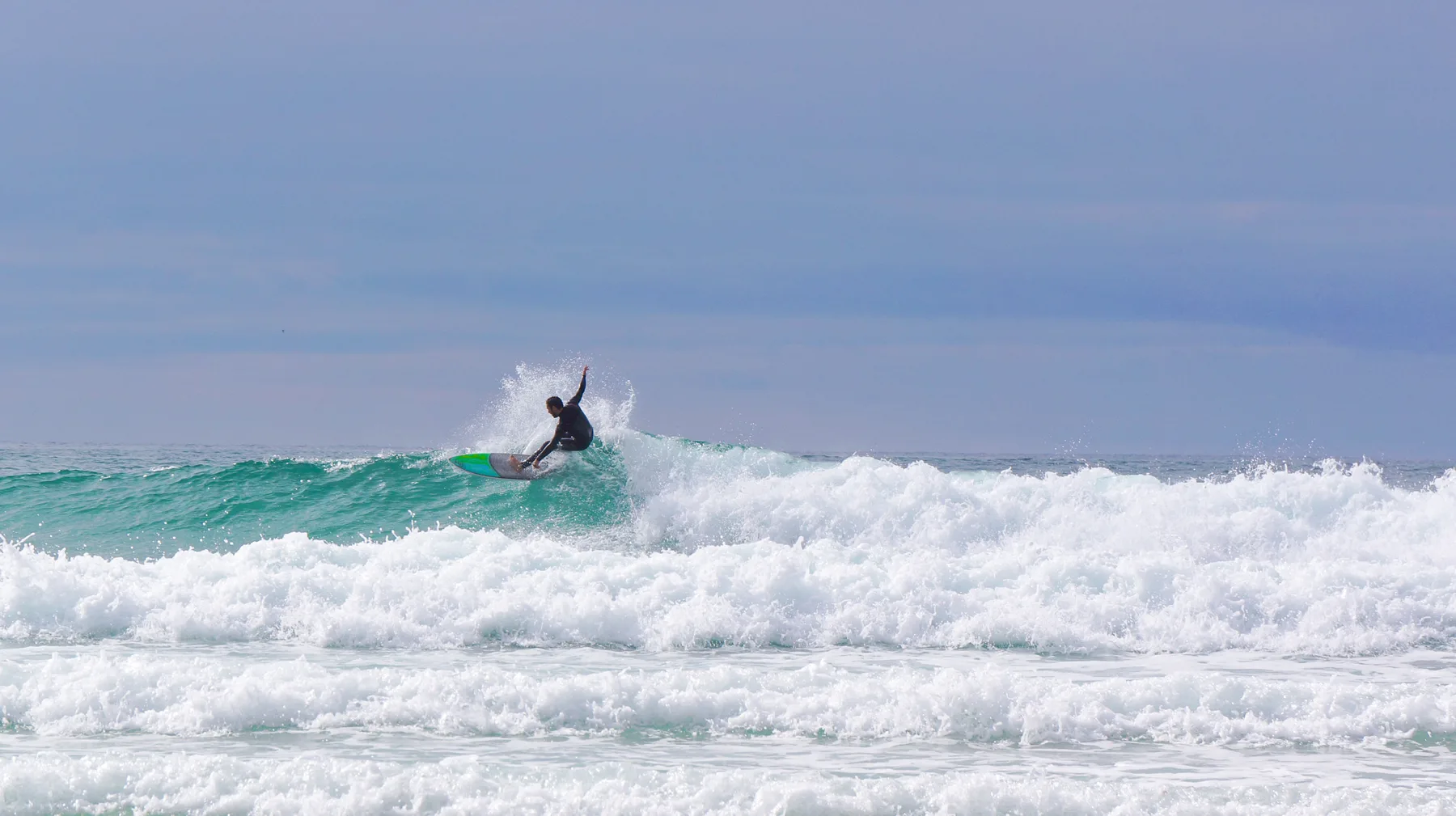 A surfer rides a breaking ocean wave beneath a clear sky.