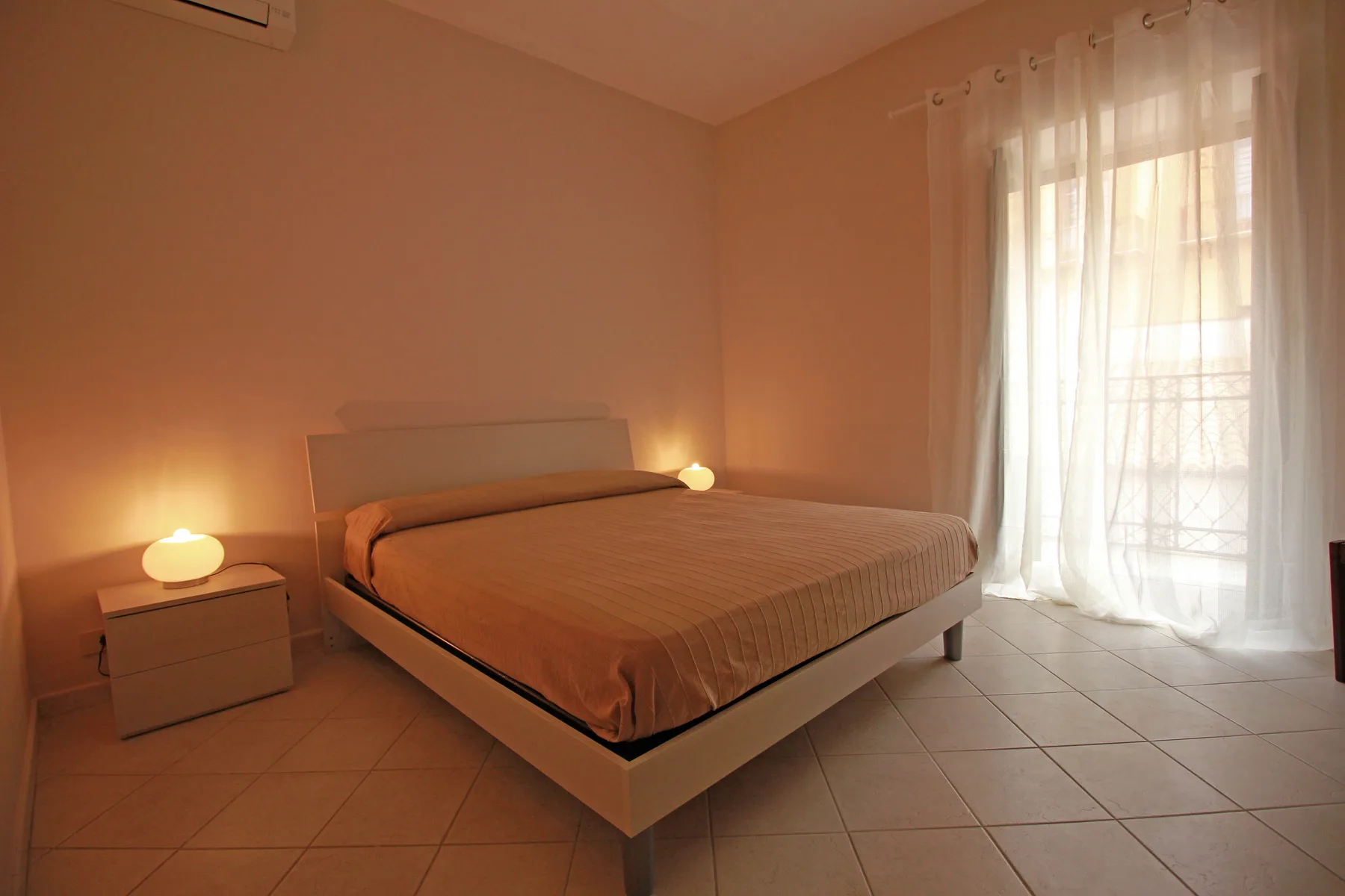 A bedroom with a bed beside a large window with sheer curtains.