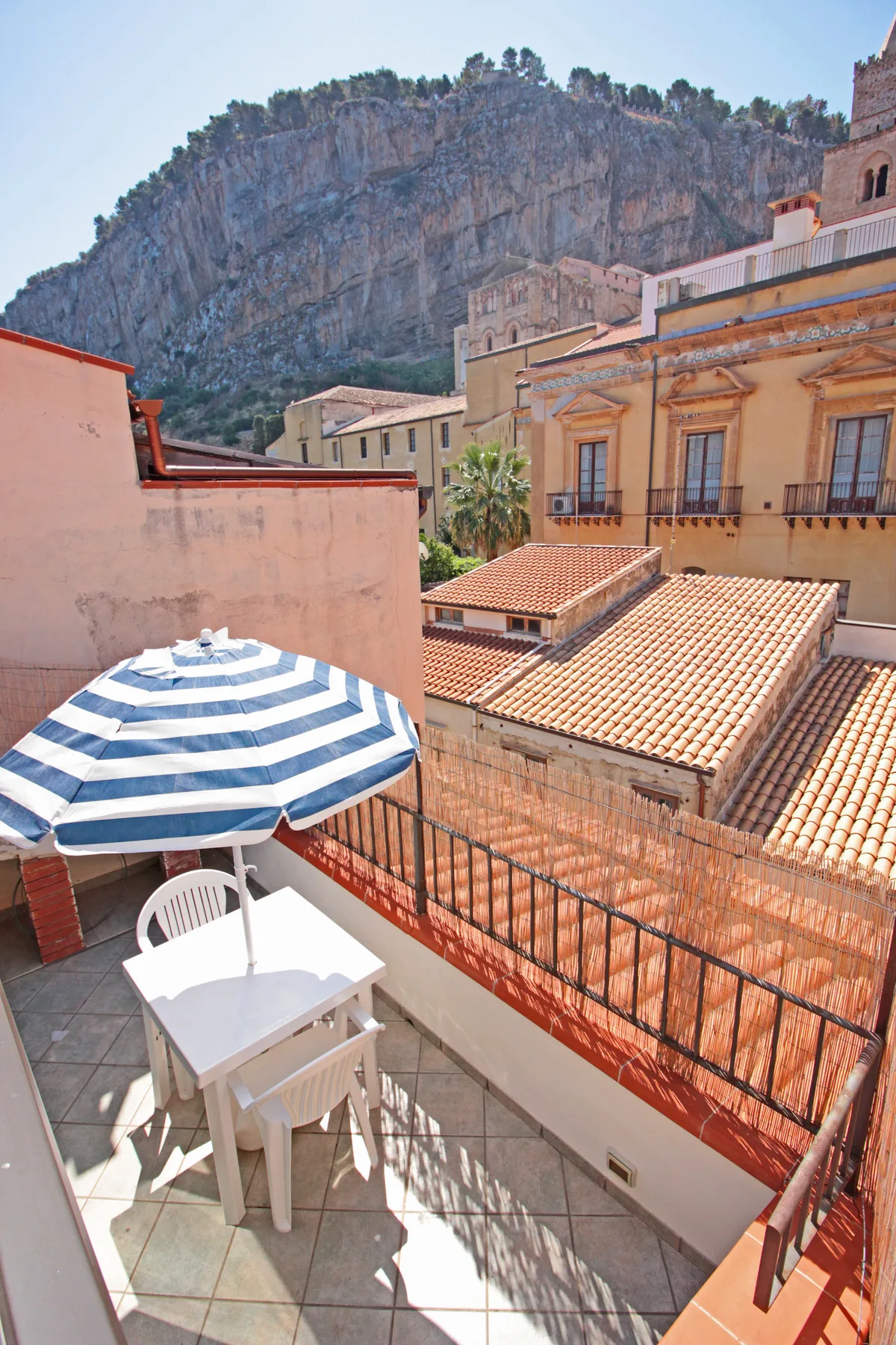 A rooftop terrace with a striped umbrella and a view of nearby buildings and a rocky mountain.