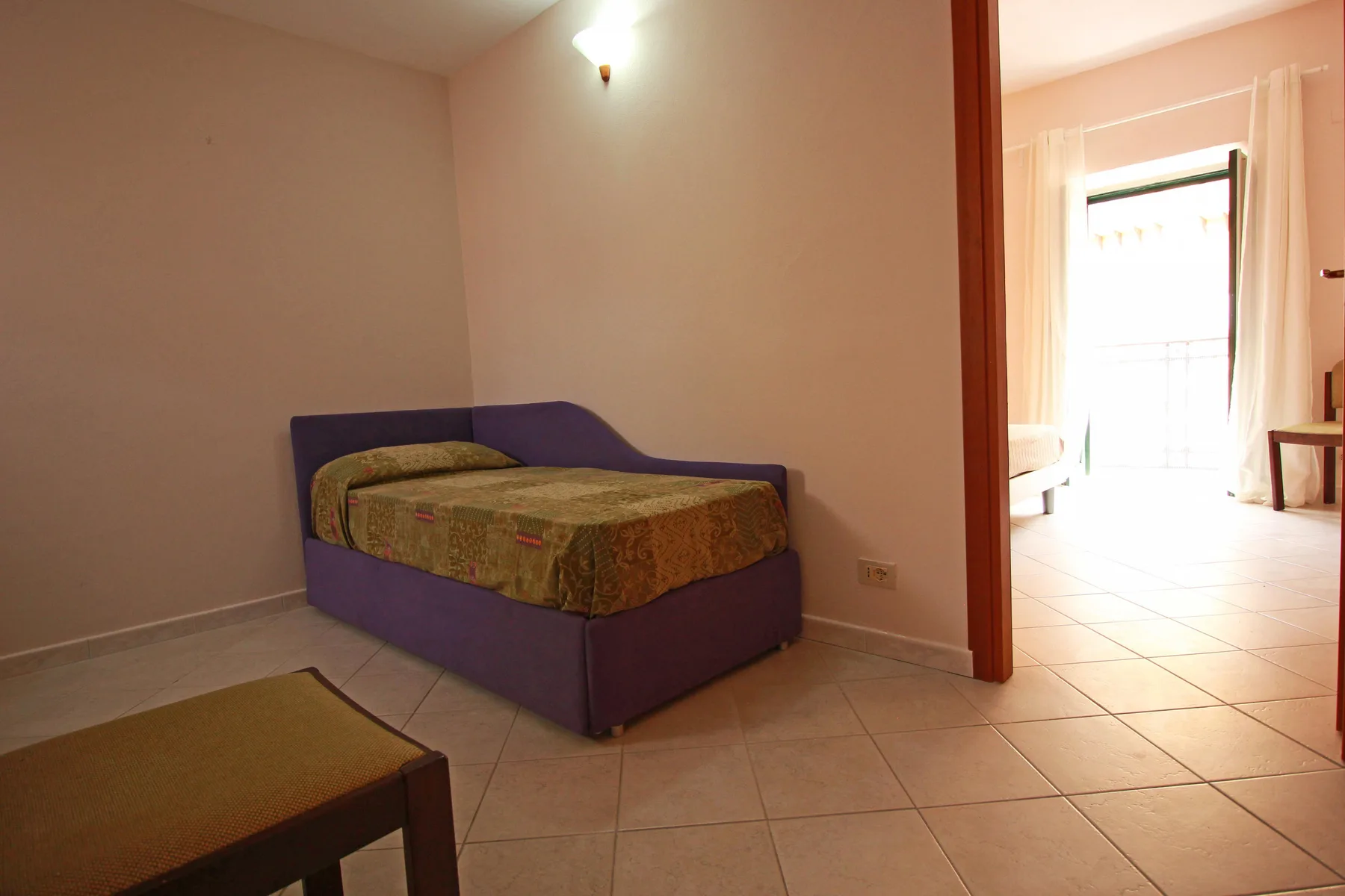 A bedroom with a single bed beside an open doorway to a bright tiled room.