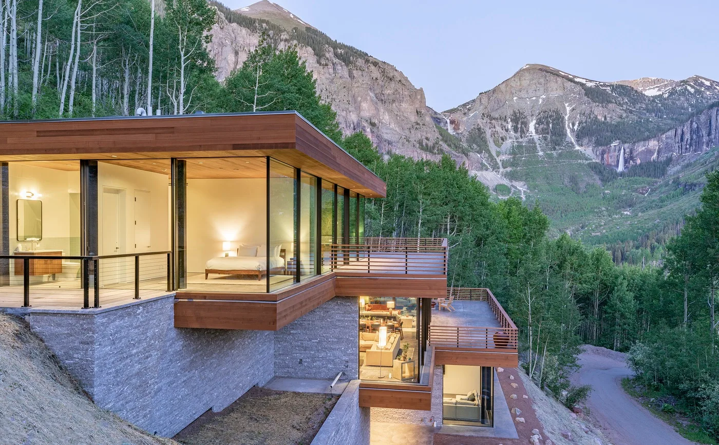 Una casa moderna con fachada de vidrio mira hacia un paisaje montañoso boscoso.